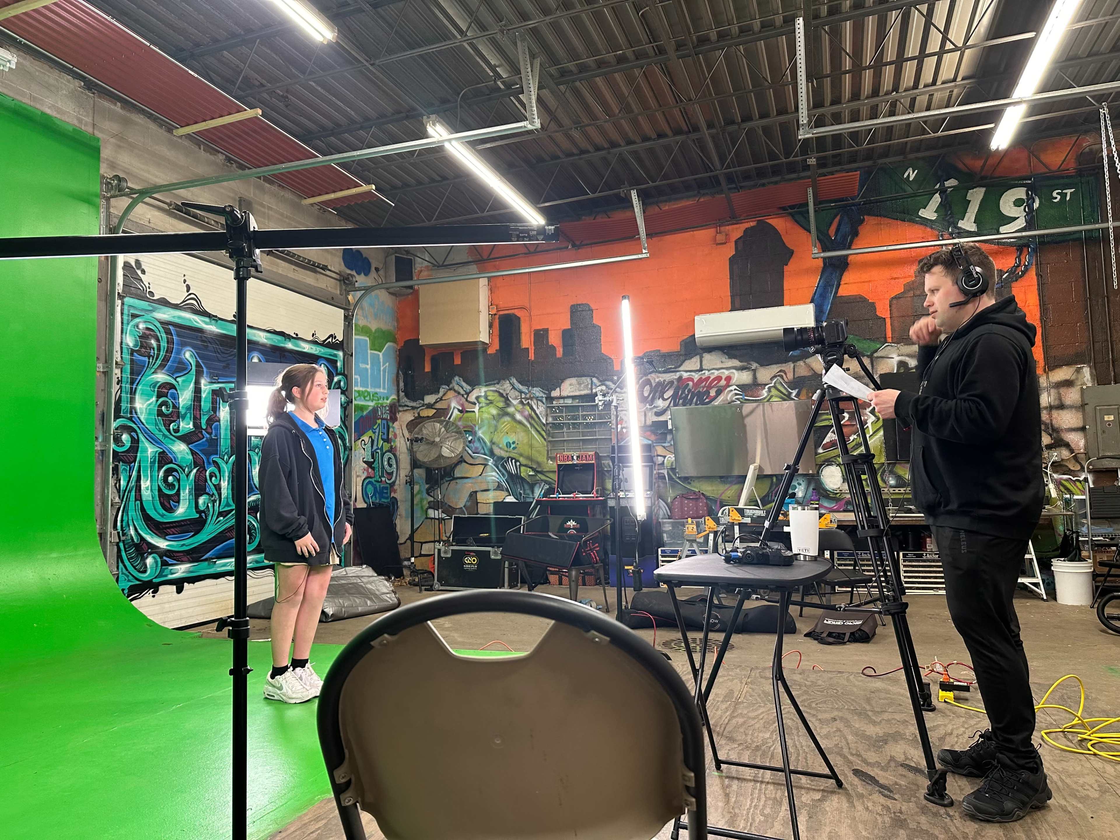 A person stands in front of a green screen while another person operates a camera in a creatively decorated studio space.