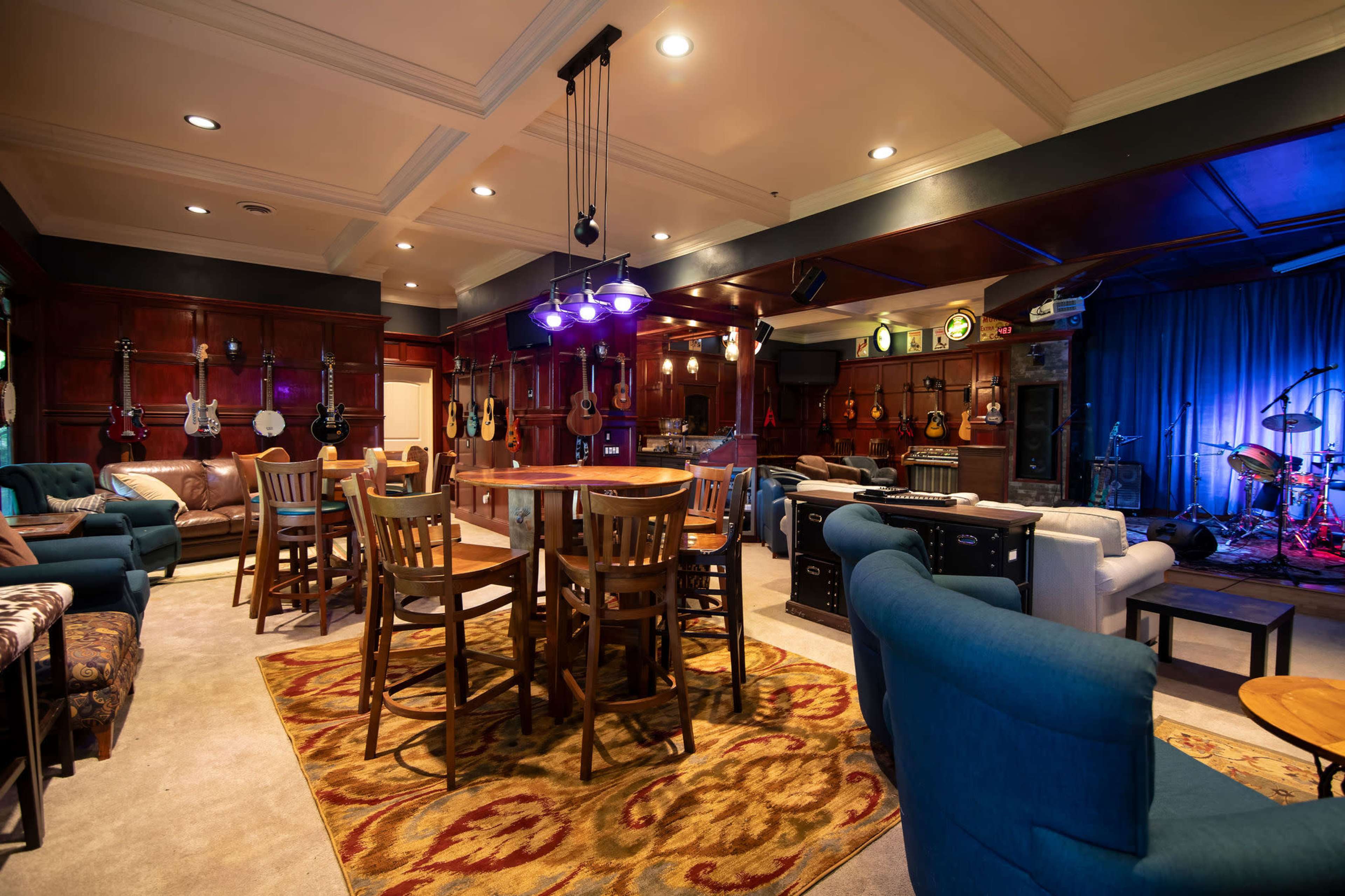 A cozy music room with wooden walls, various guitars displayed, seating areas, and a small stage illuminated in blue light.