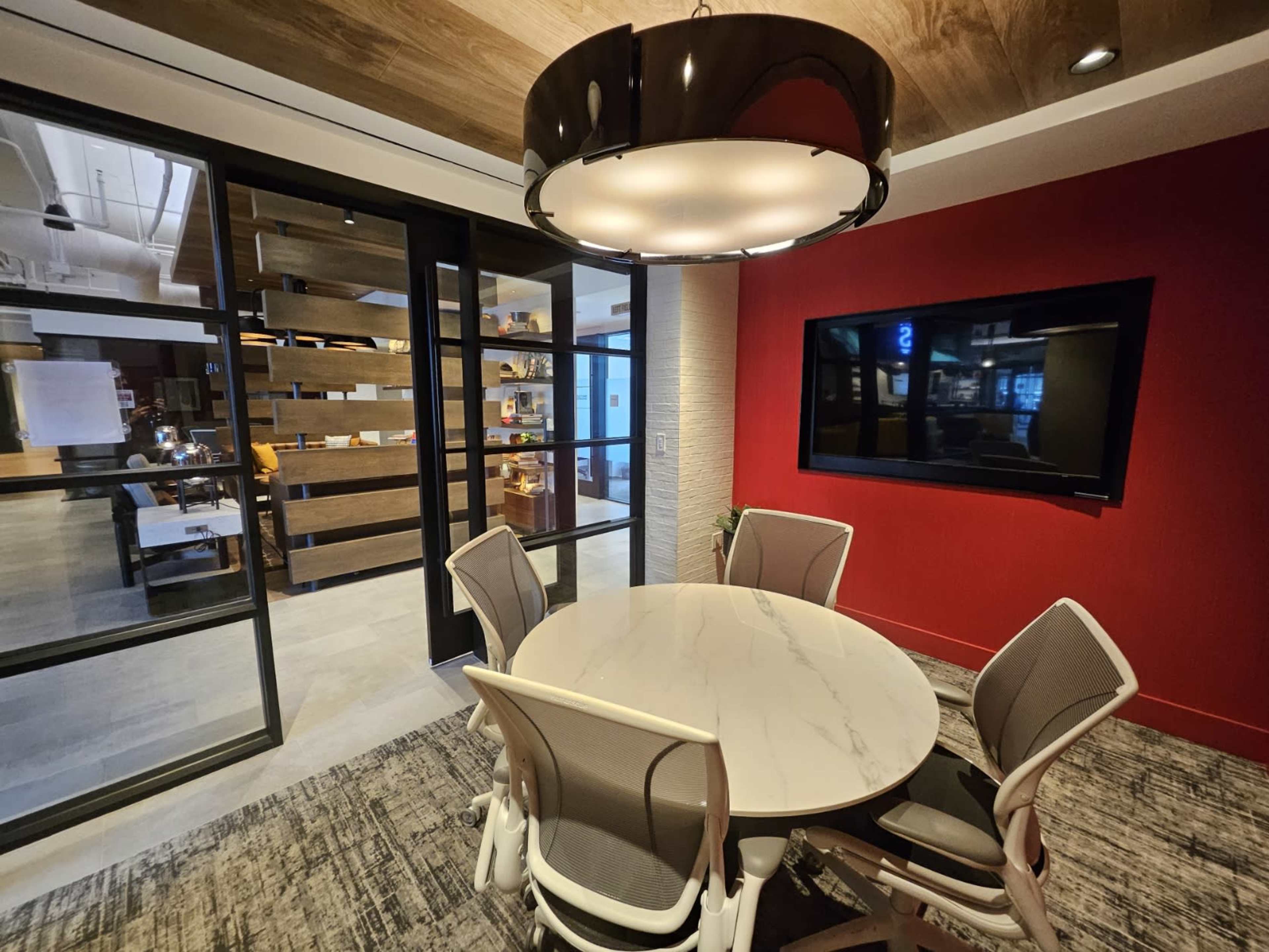 A conference room features a round marble table surrounded by chairs, with a large screen and a red accent wall in the background.