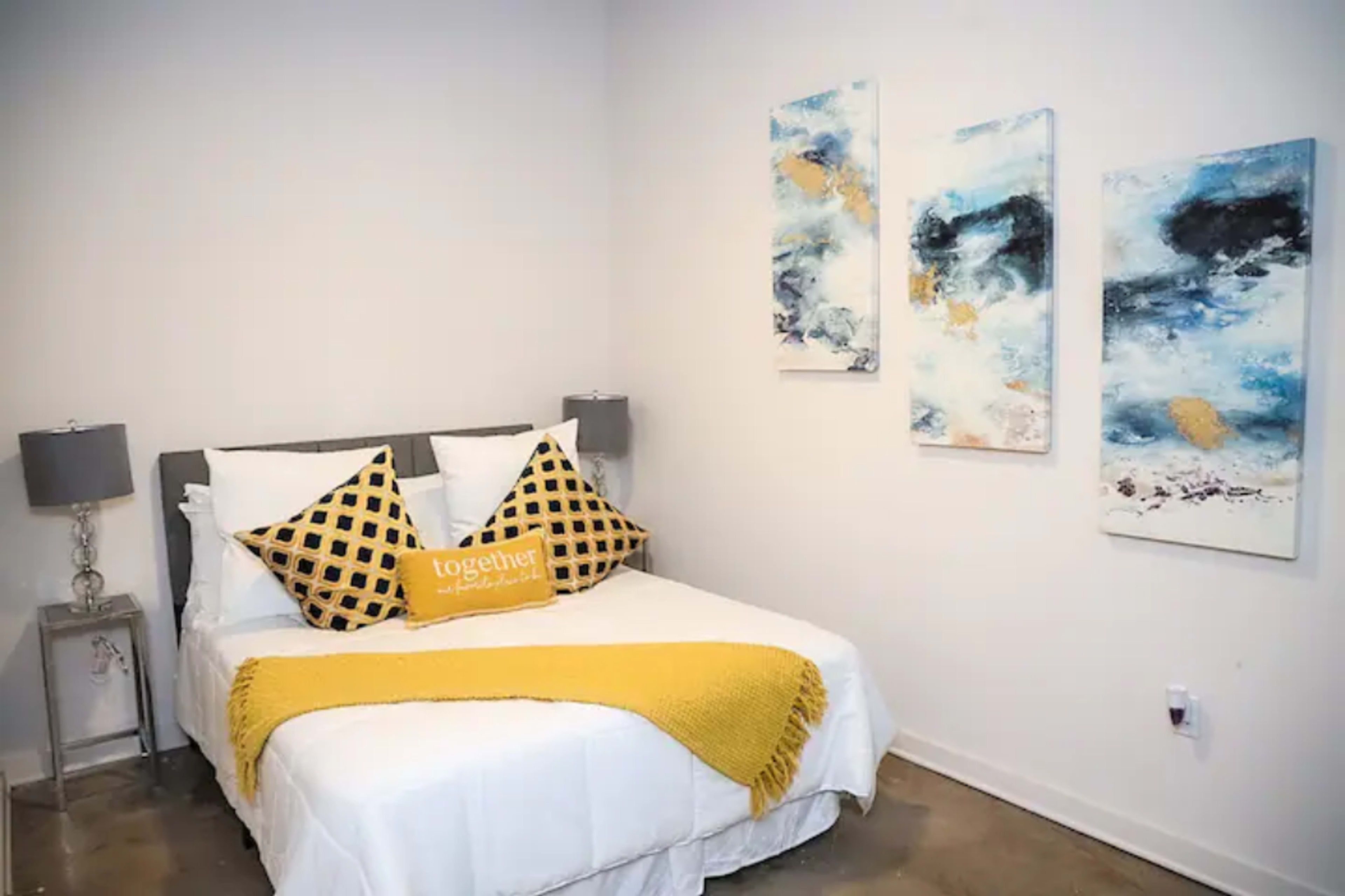 A small bedroom features a bed with a white comforter, yellow accent pillows, and art pieces on the wall.