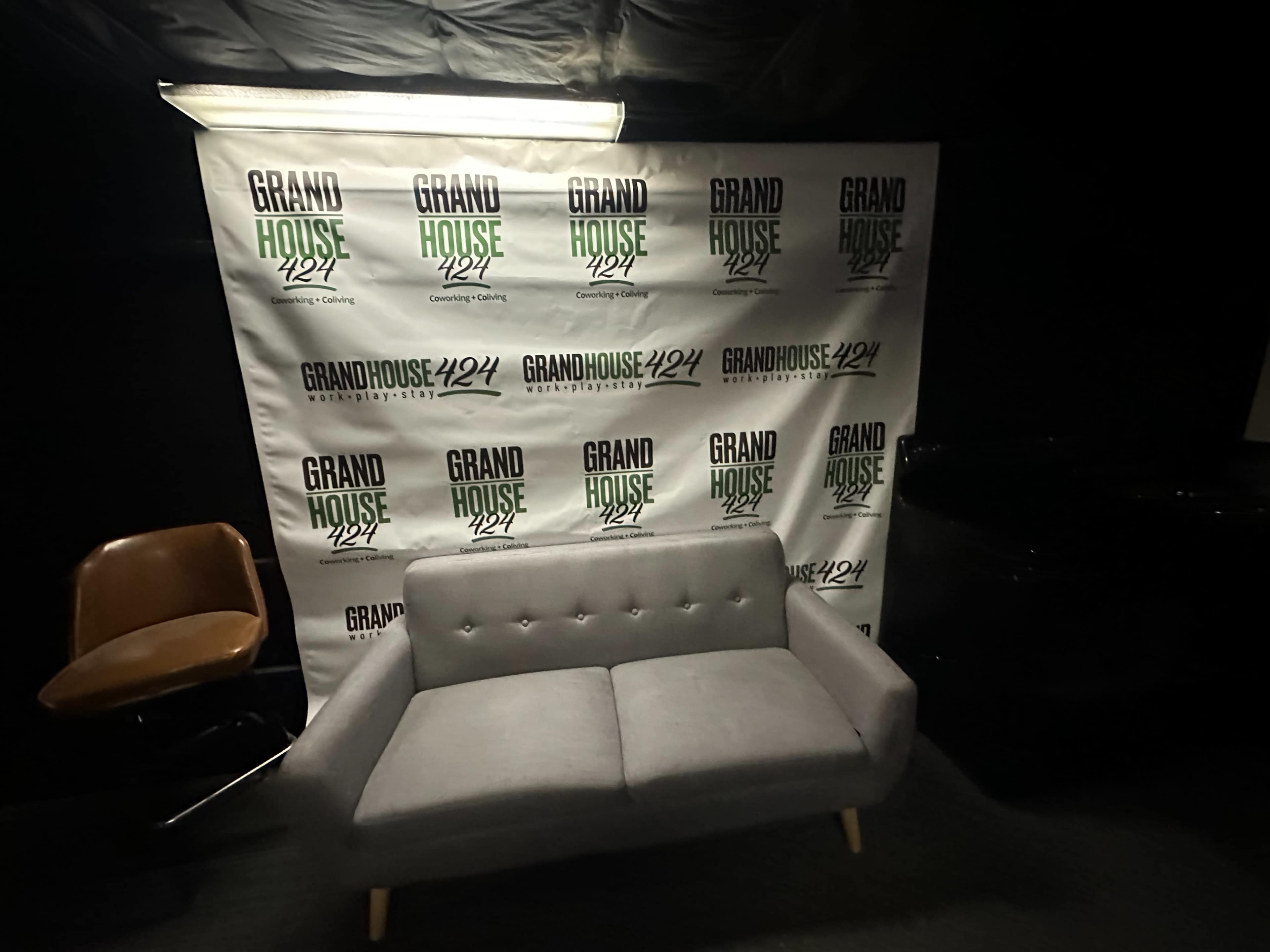 The image shows a gray sofa in front of a banner featuring the text "GRAND HOUSE 424" with a repeating pattern.