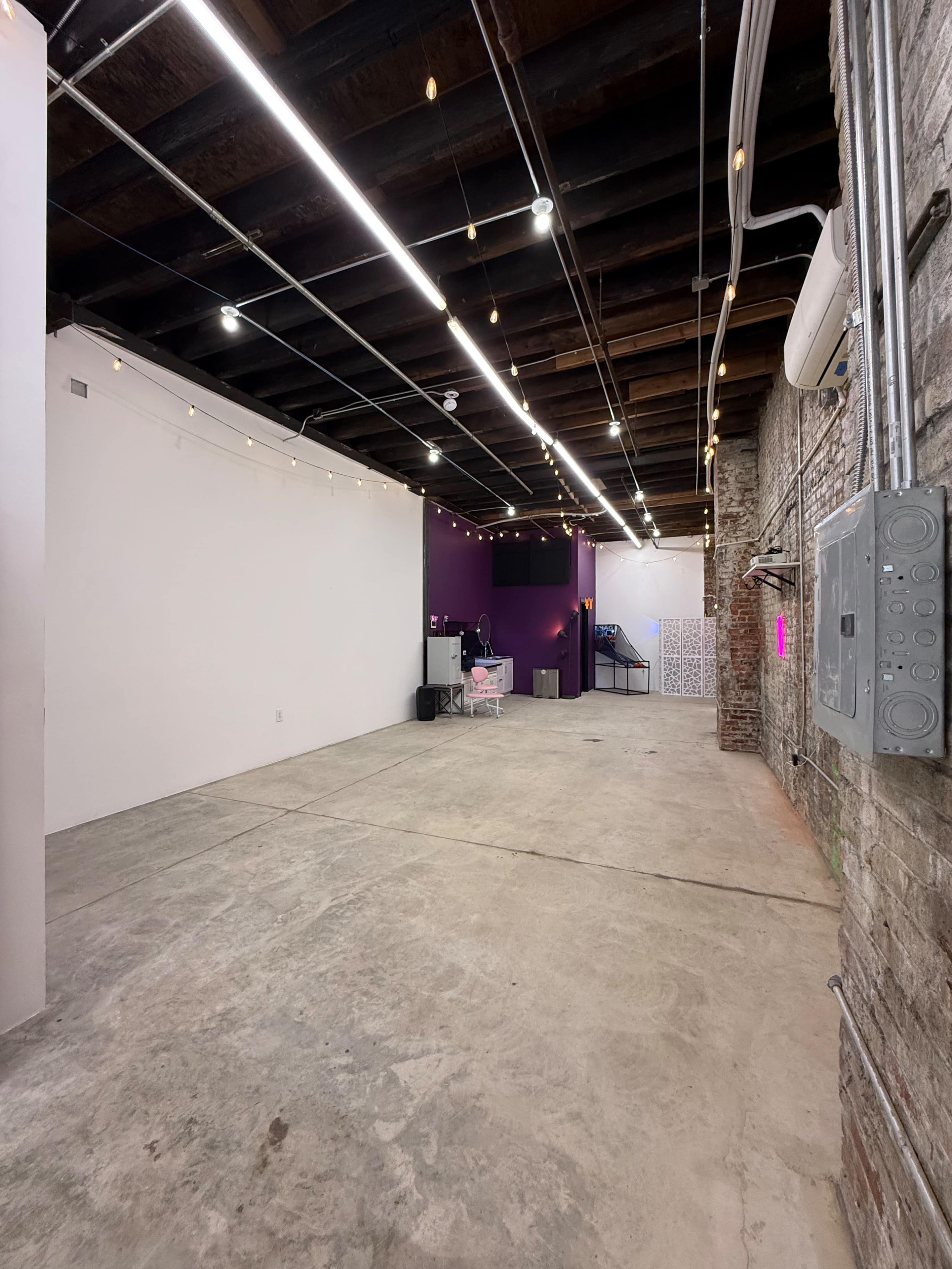 The image shows a spacious interior with exposed brick walls, concrete flooring, and a mix of white and purple areas, illuminated by overhead lights and decorative string lights.