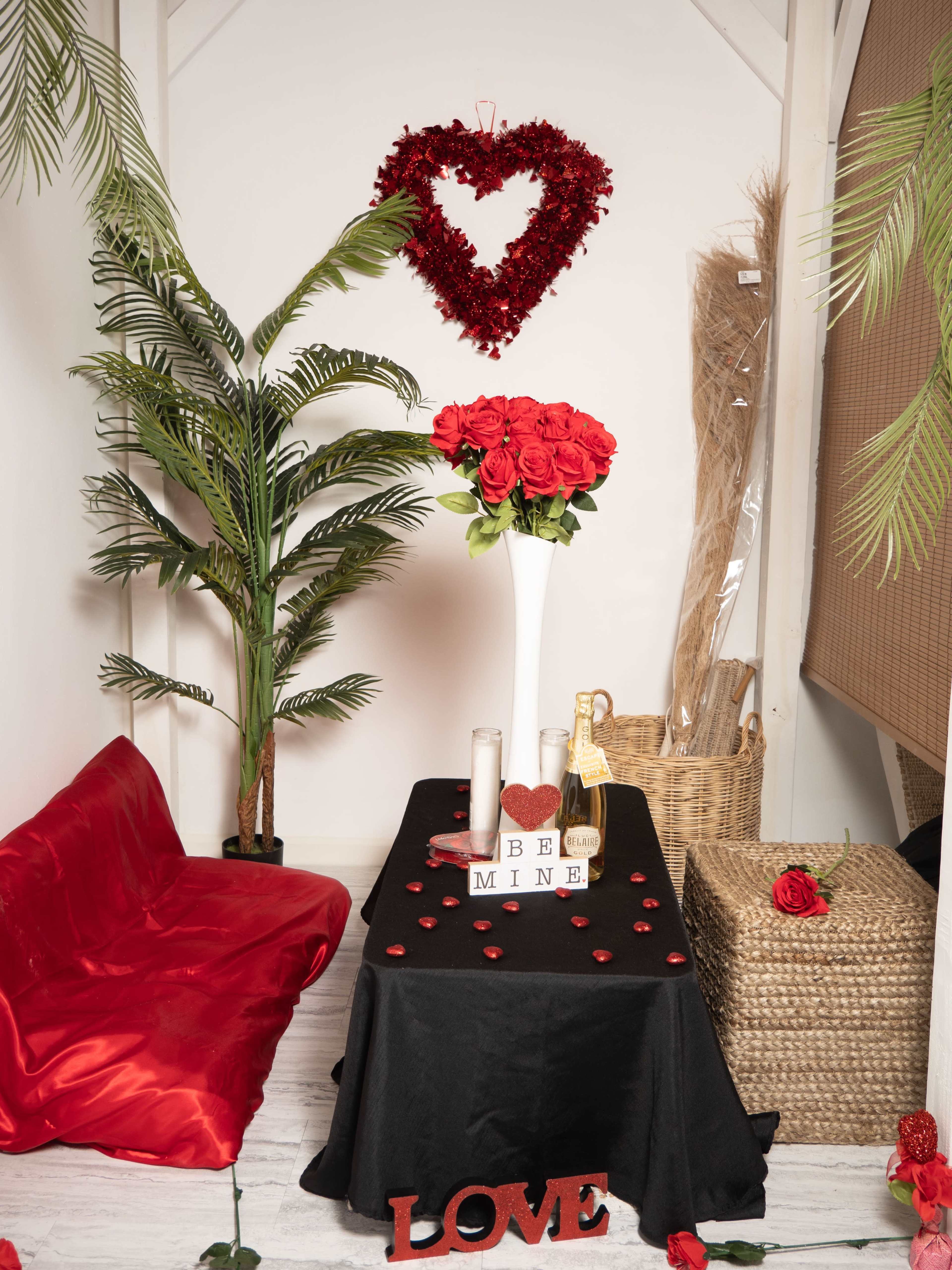 The scene features a table draped in black cloth adorned with candles, rose petals, and a bottle of champagne, set against a backdrop of decorative plants and a heart-shaped wreath.