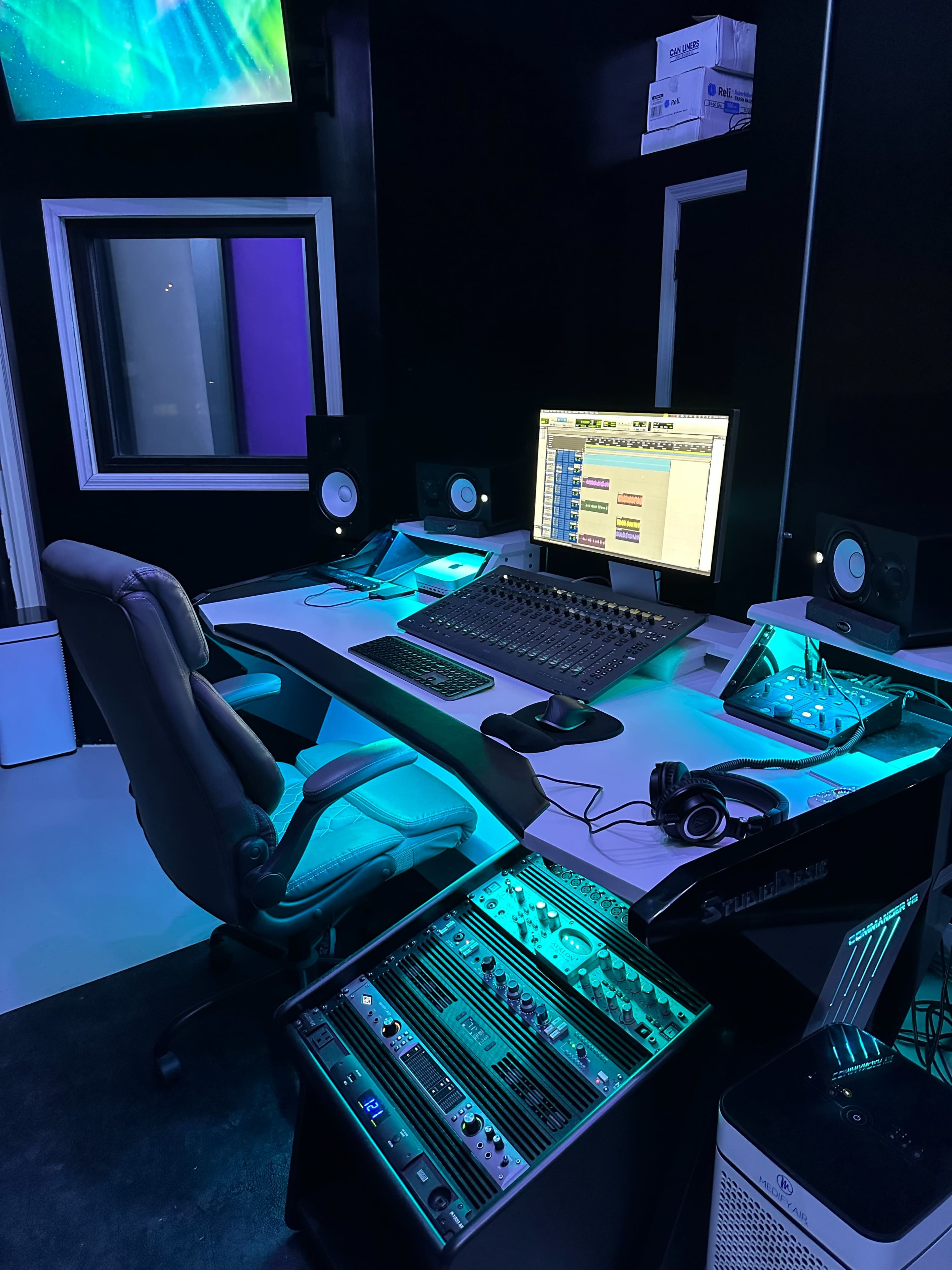 A modern recording studio is equipped with a computer, audio interface, monitors, and a comfortable chair, illuminated by soft blue lighting.