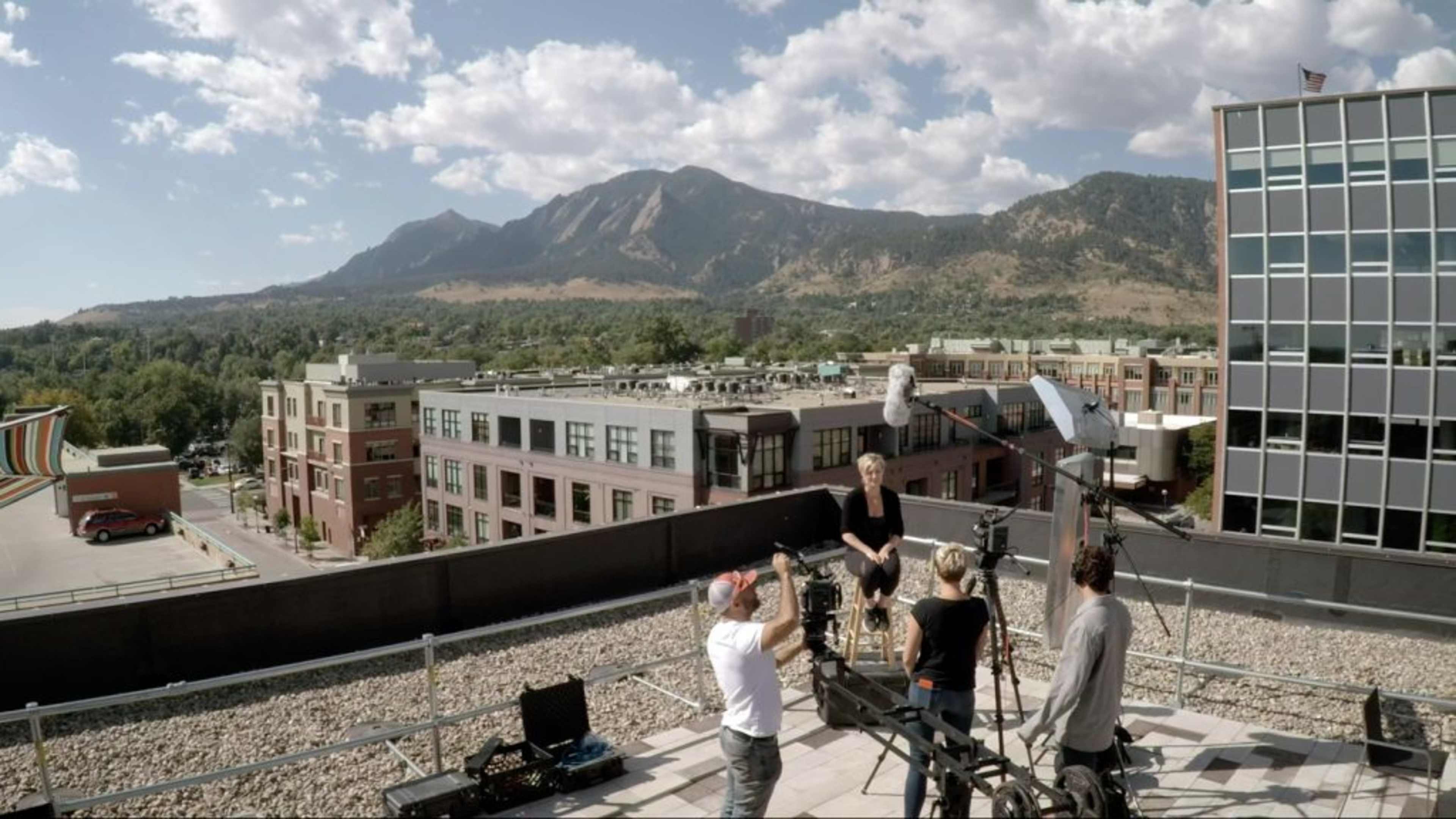 A film crew is capturing footage on a rooftop with a backdrop of mountains and urban buildings.