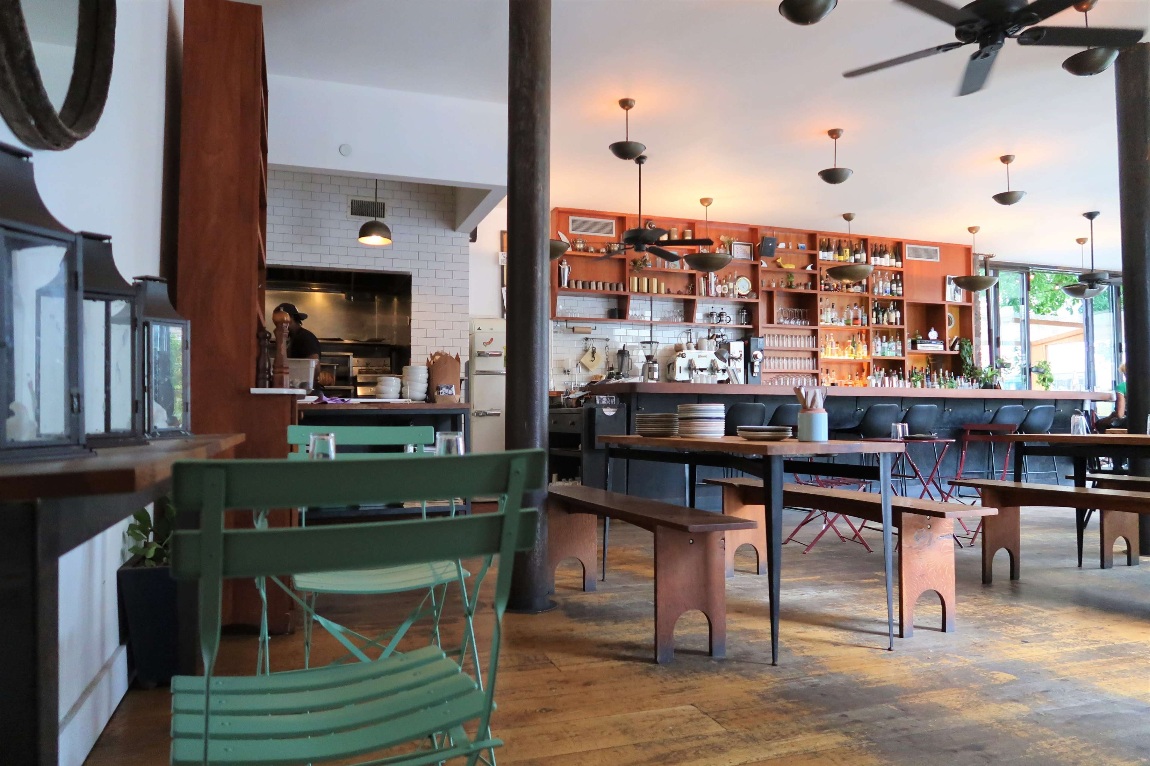 The interior of a restaurant featuring wooden floors, a bar stocked with bottles, and a kitchen area visible in the background.