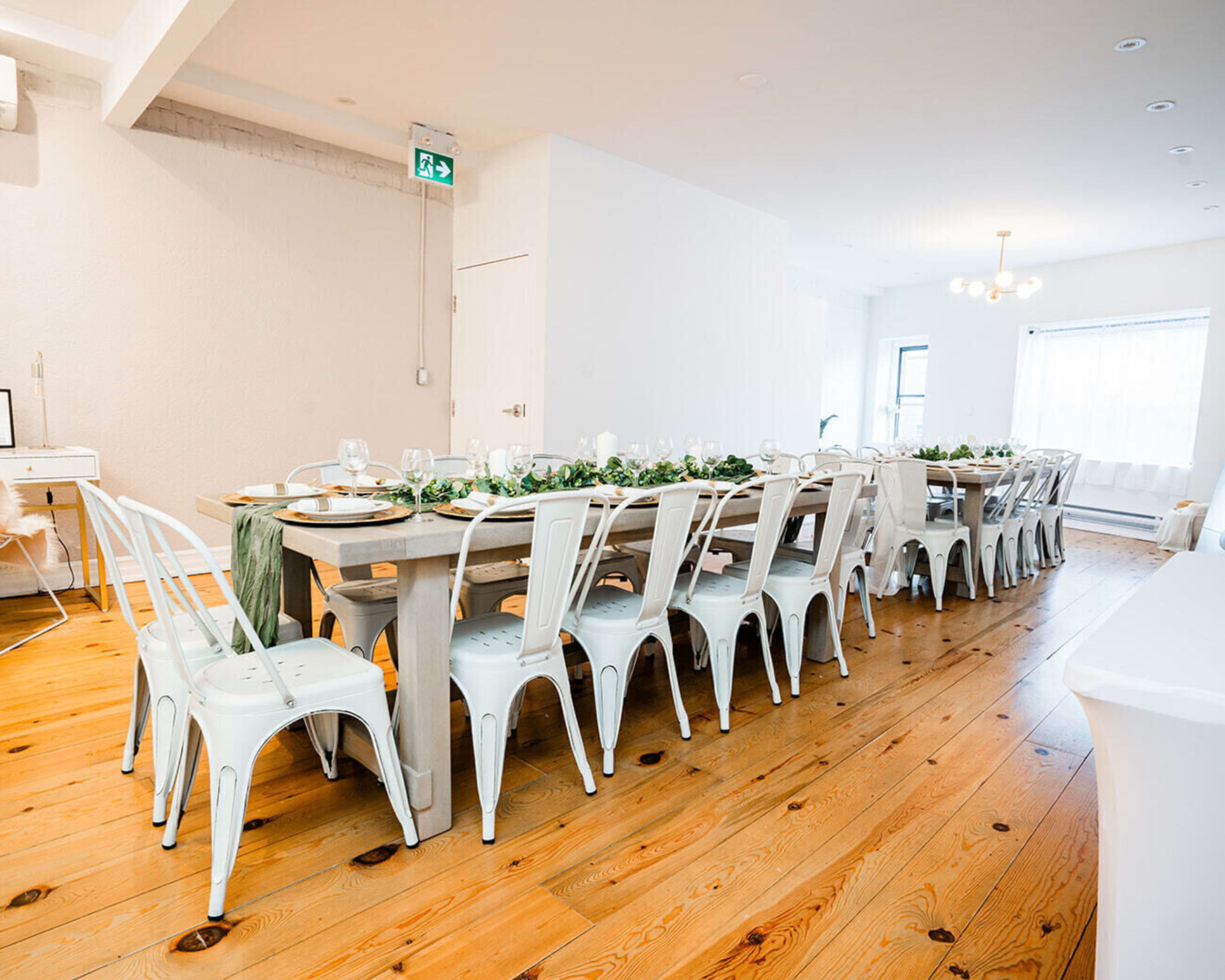 A long wooden dining table set for a meal is arranged in a spacious room with white walls and wooden flooring.