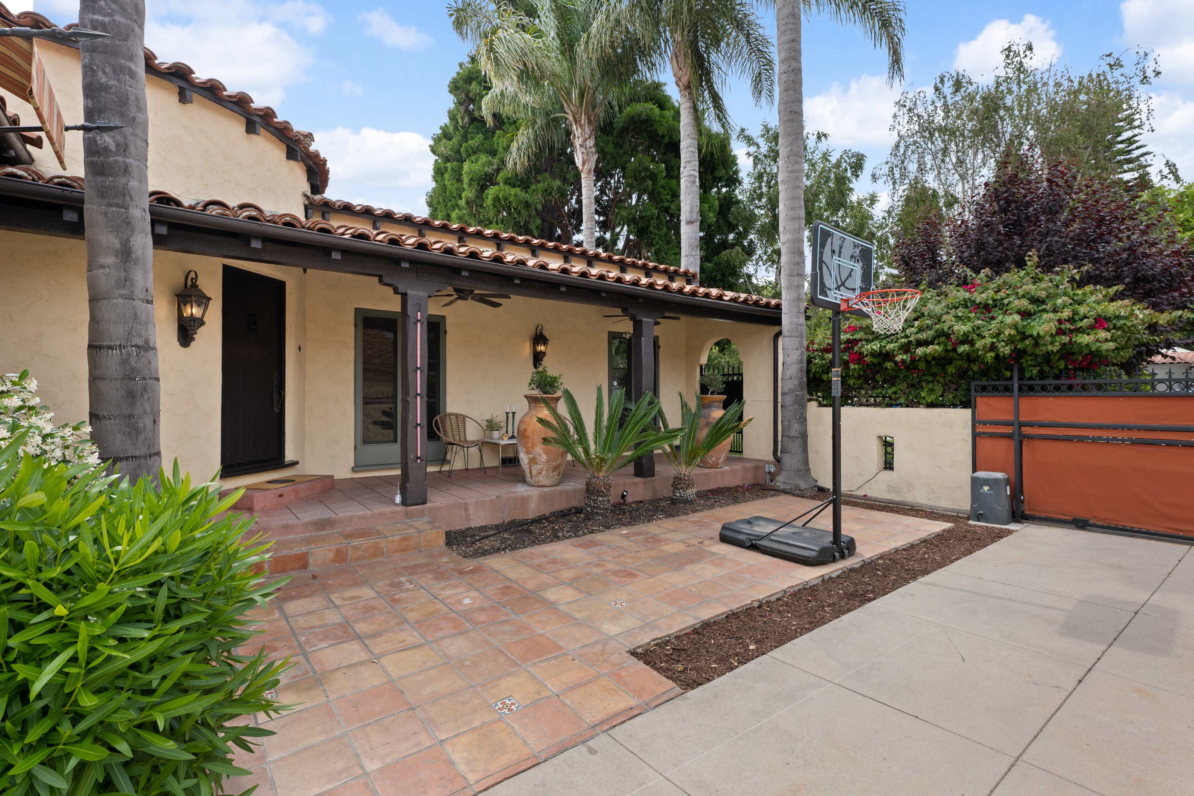 The image shows a patio area of a house featuring a basketball hoop, surrounded by potted plants and palm trees.