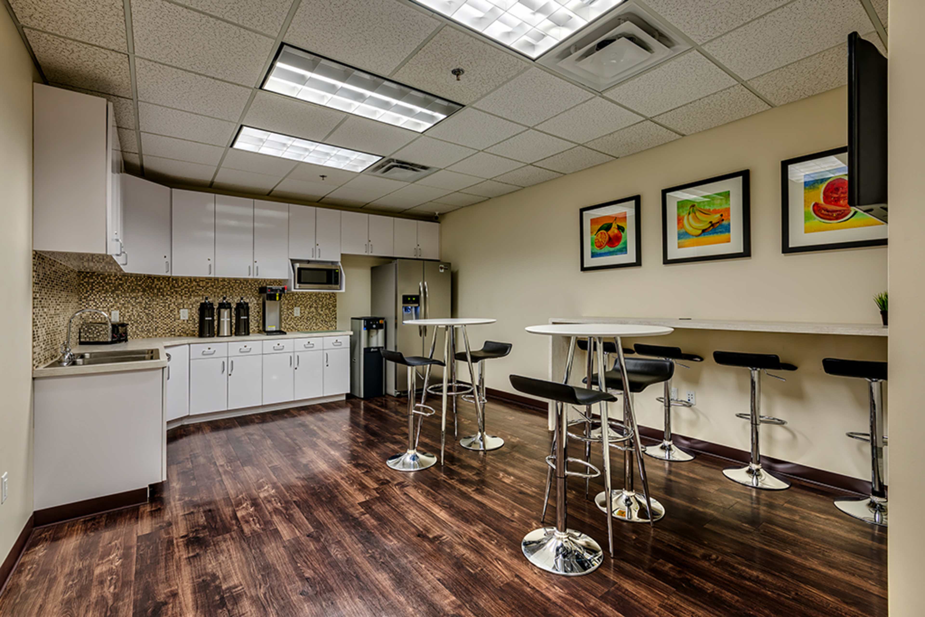 A modern break room with a kitchenette, featuring white cabinets, a microwave, coffee machine, and bar-style seating.