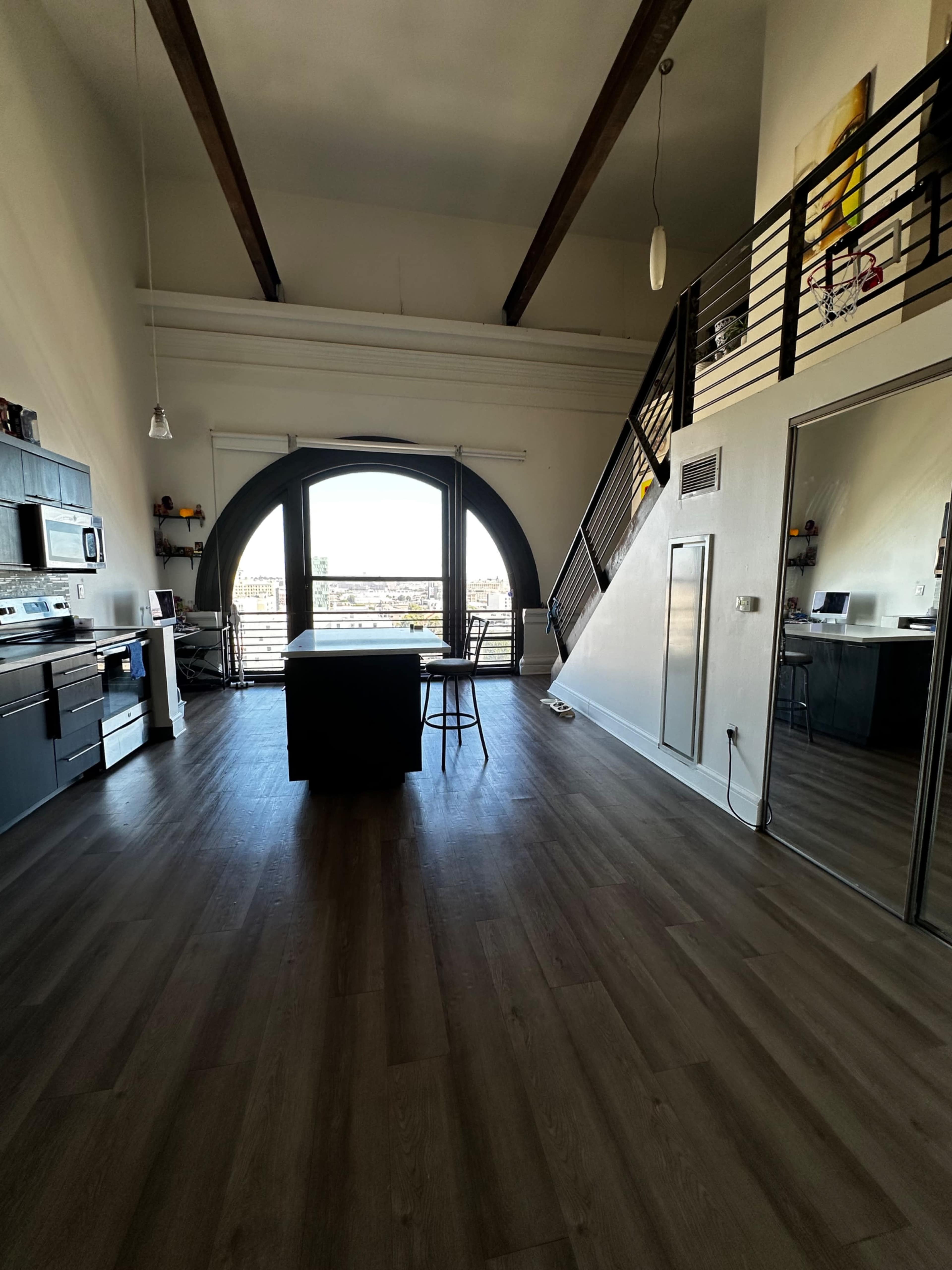 The image shows a spacious loft-style apartment with a modern kitchen, a large arched window, and a staircase leading to an upper level.
