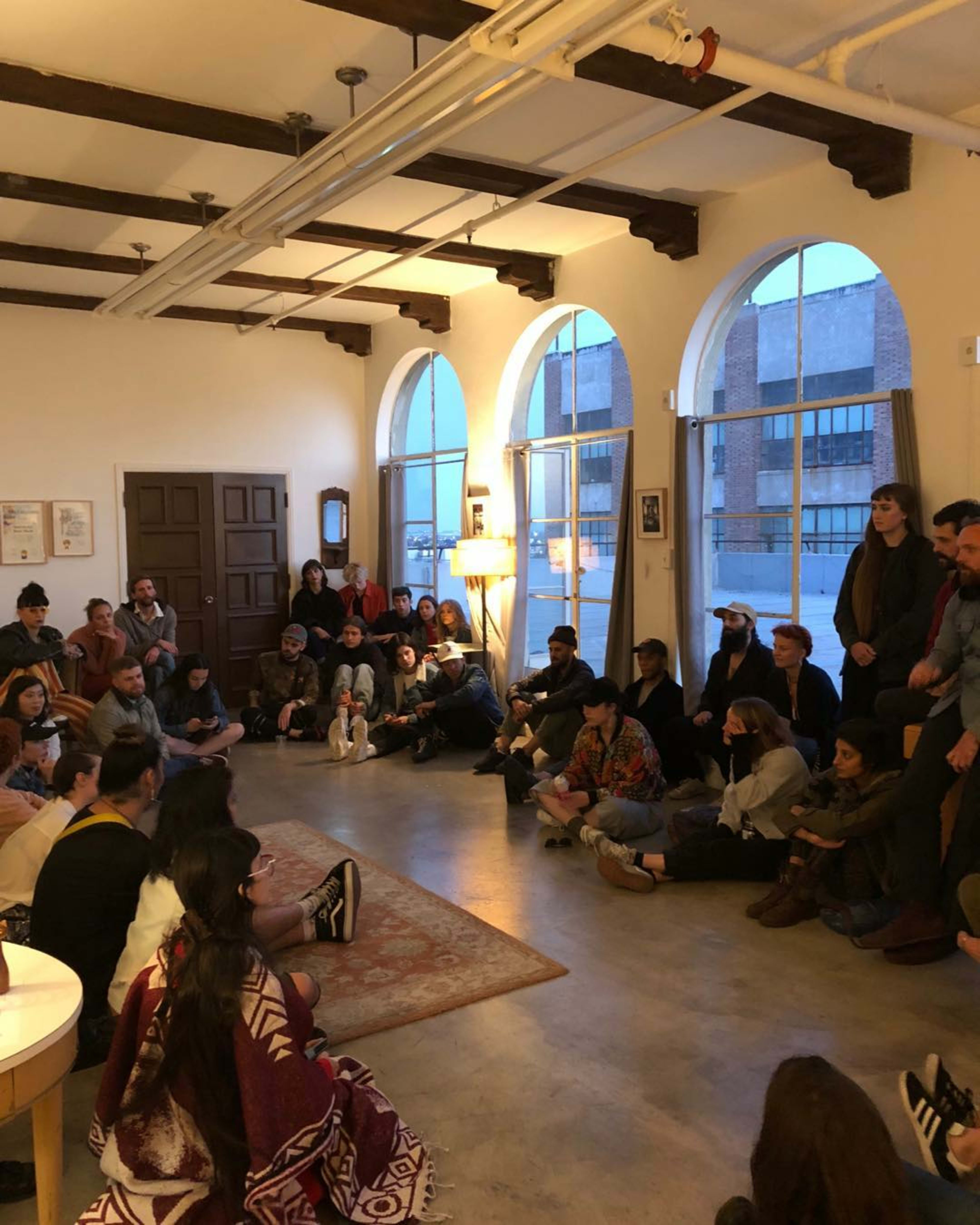 A group of people sits on the floor of a room with large arched windows, engaged in a gathering around a low table.