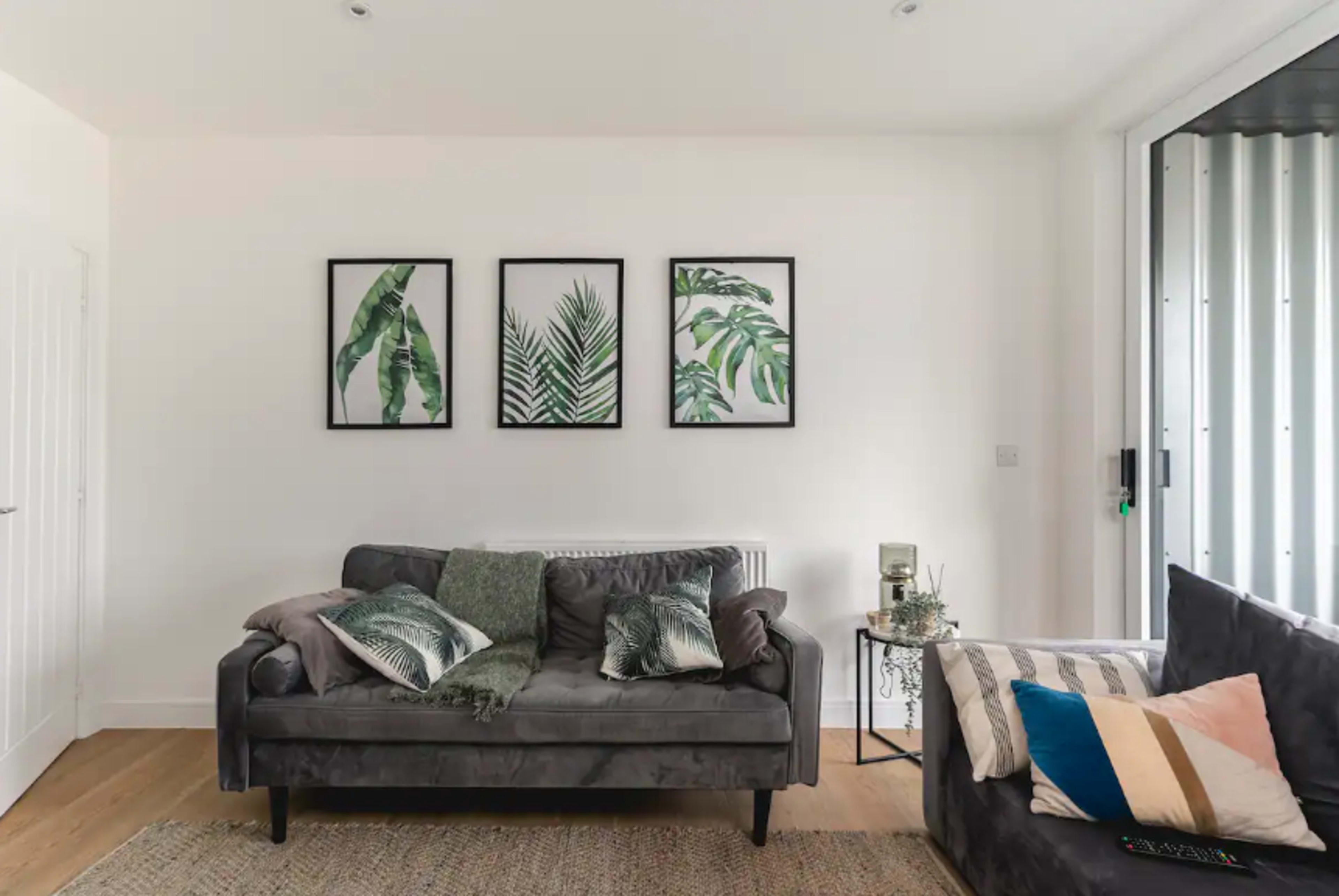 The image shows a living room with a grey sofa, a dark couch, and three framed botanical prints on the wall.