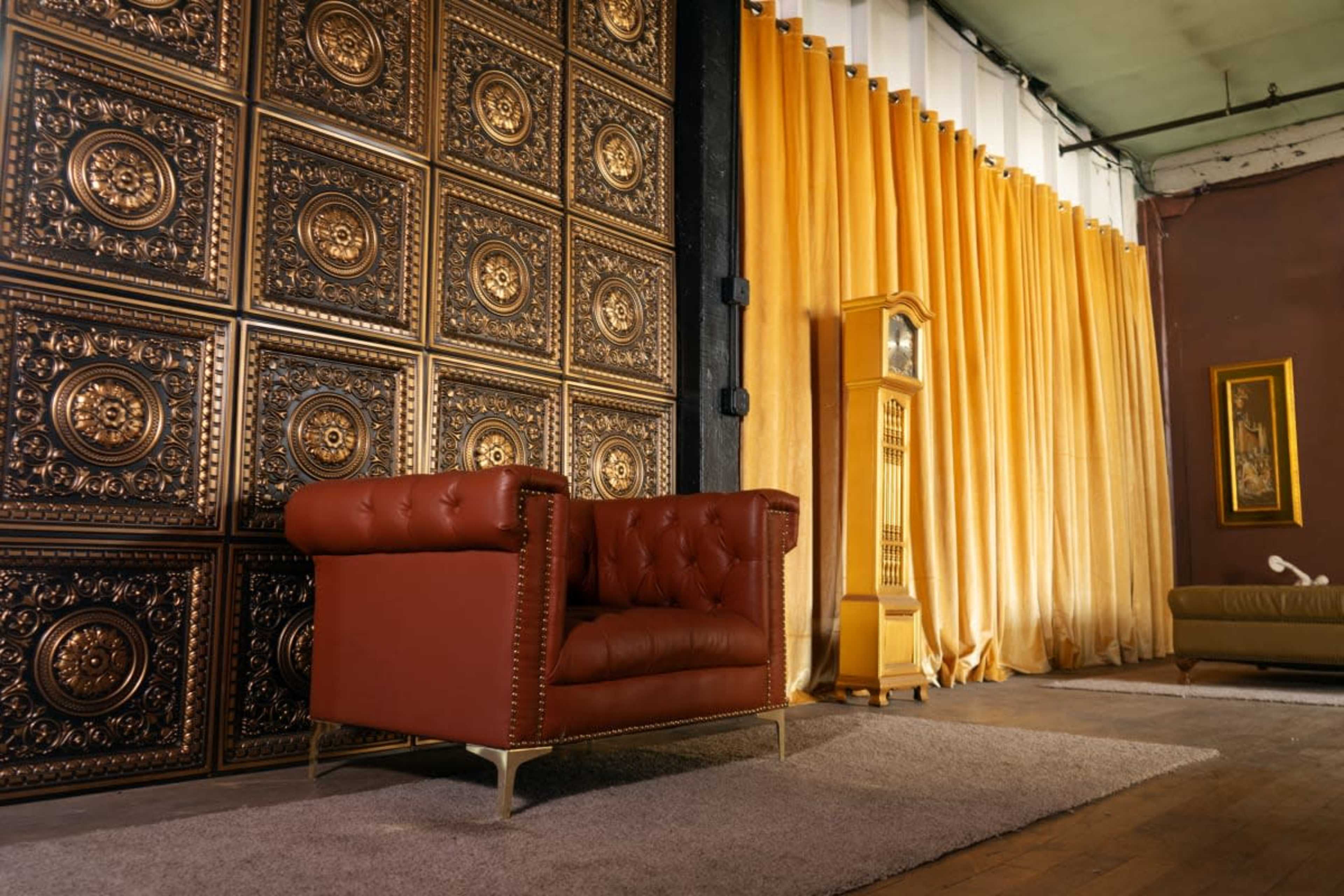 A brown leather armchair is positioned on a rug in front of a wall with ornate gold panels and golden drapes.