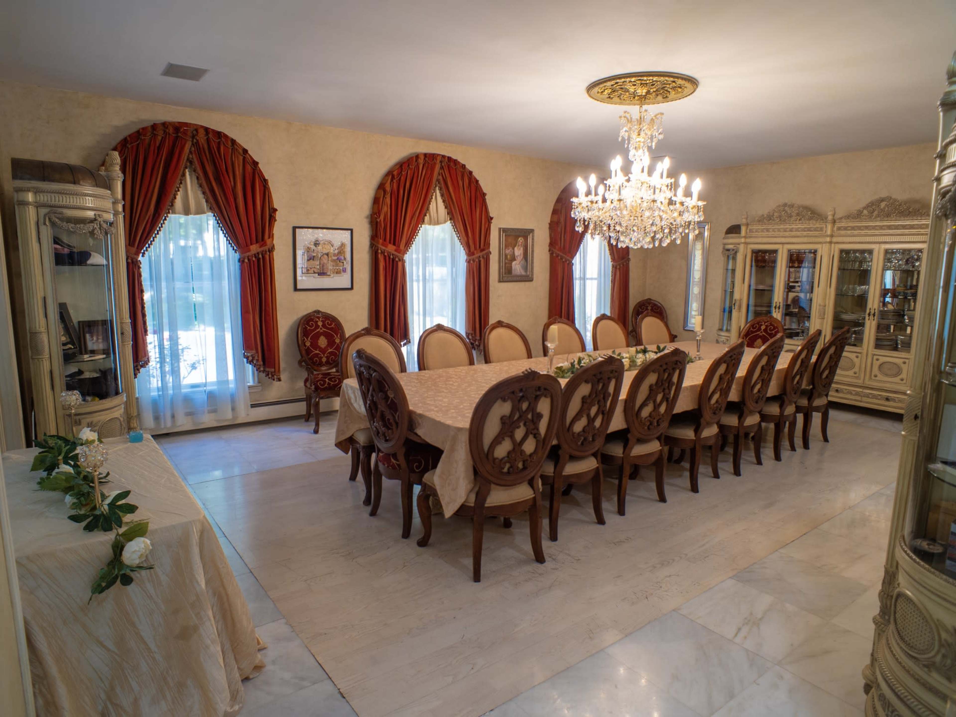 The image shows an elegantly furnished dining room featuring a large rectangular table surrounded by twelve ornate chairs, with a chandelier overhead and decorative curtains at the windows.