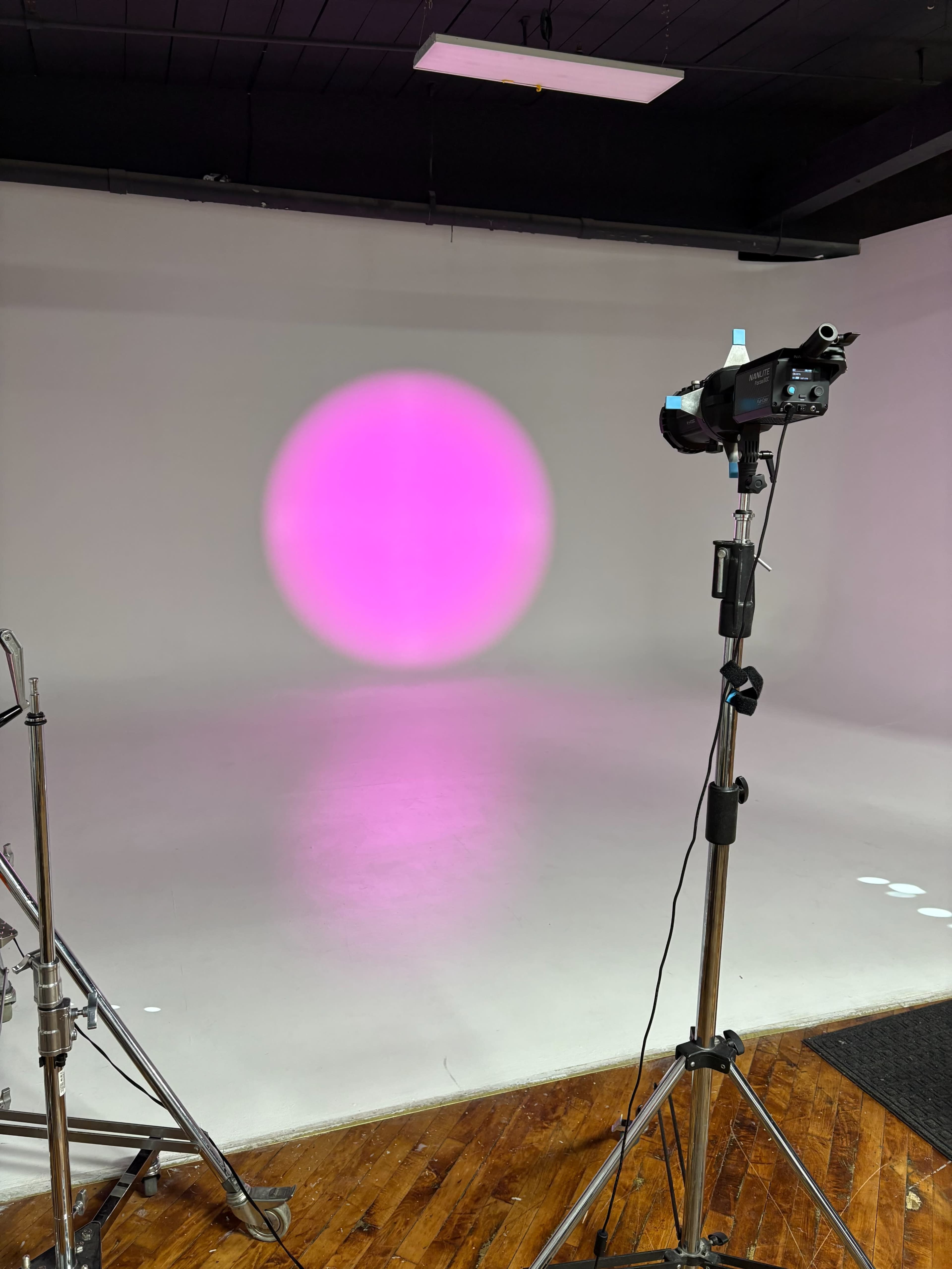 A photography studio features a pink circular light projection on a white wall, with a lighting stand positioned nearby.