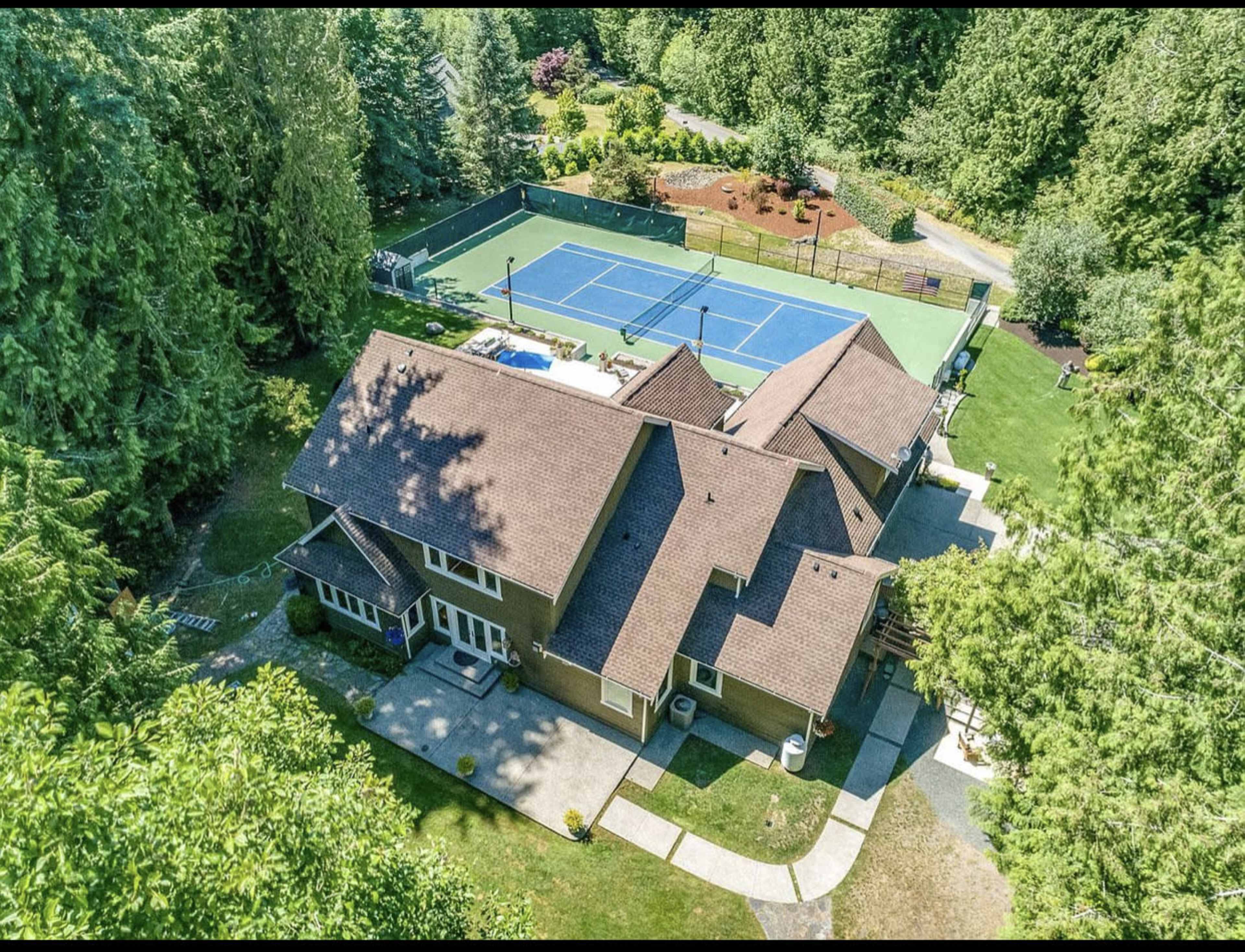 An aerial view of a large house surrounded by trees, featuring a tennis court and a swimming pool in the backyard.