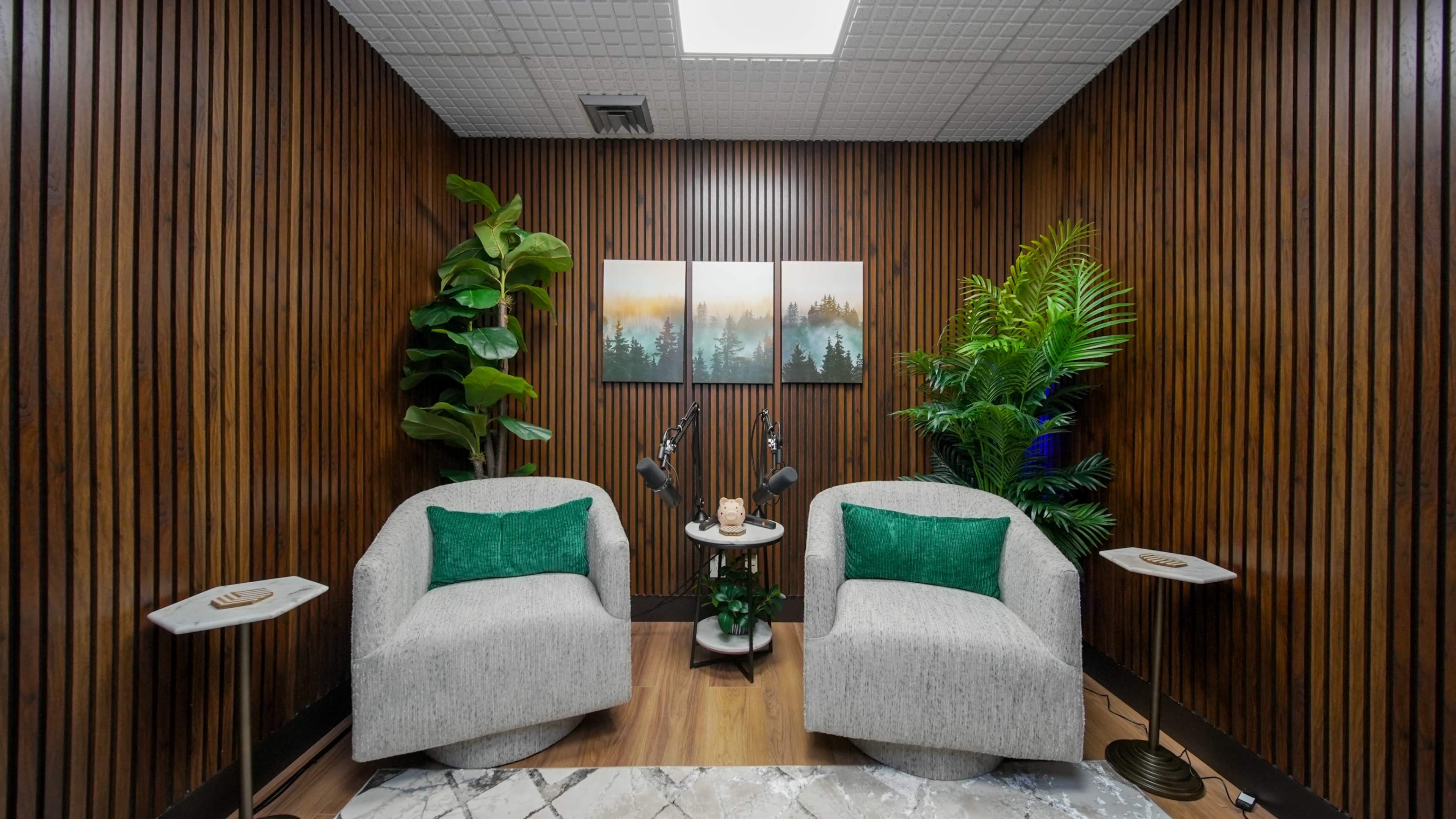 The image shows a cozy seating area with two light gray chairs, small side tables, and decorative plants against a wooden-paneled wall.