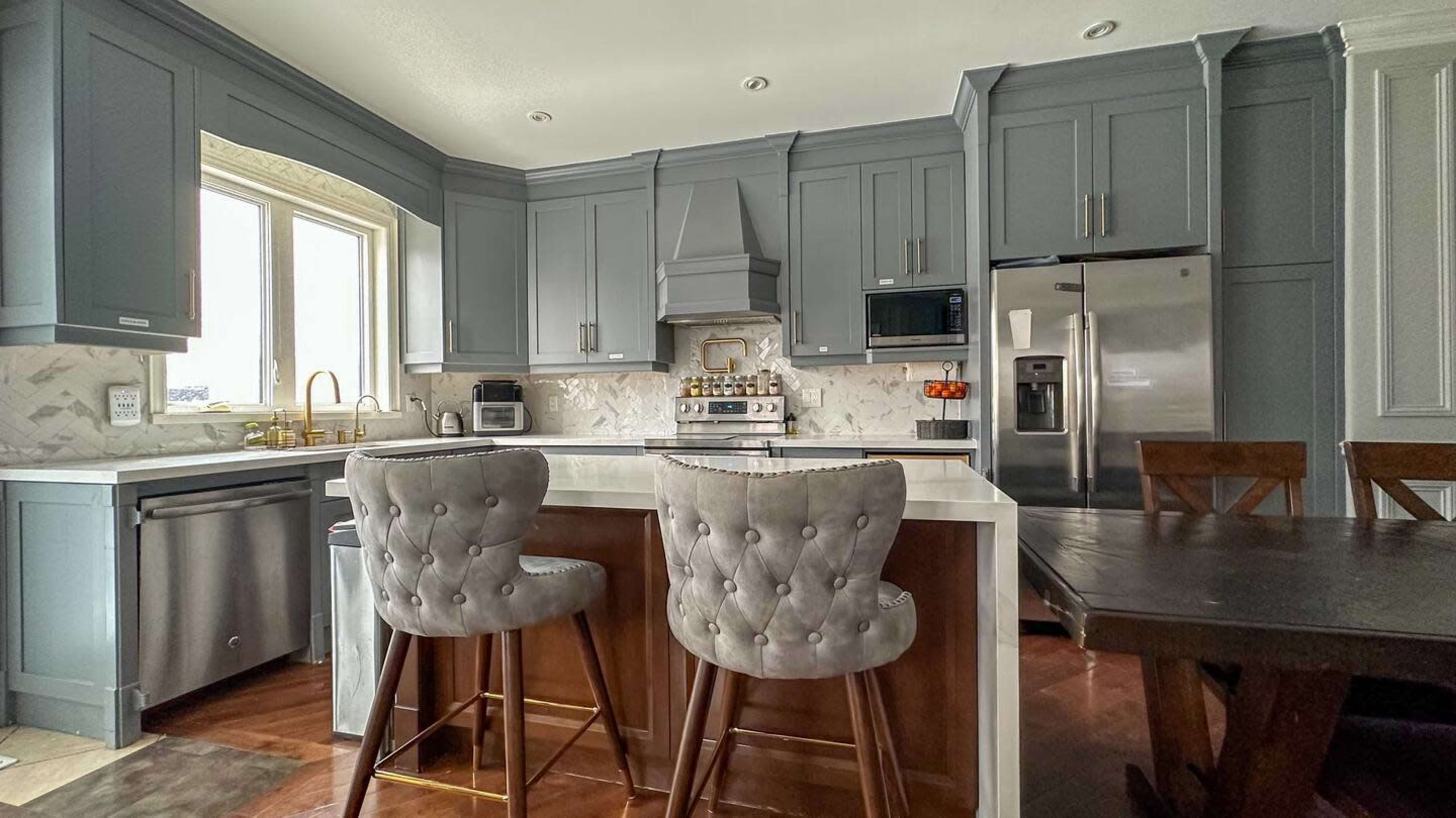 A modern kitchen features gray cabinetry, a white countertop, and two upholstered bar stools at a central island.