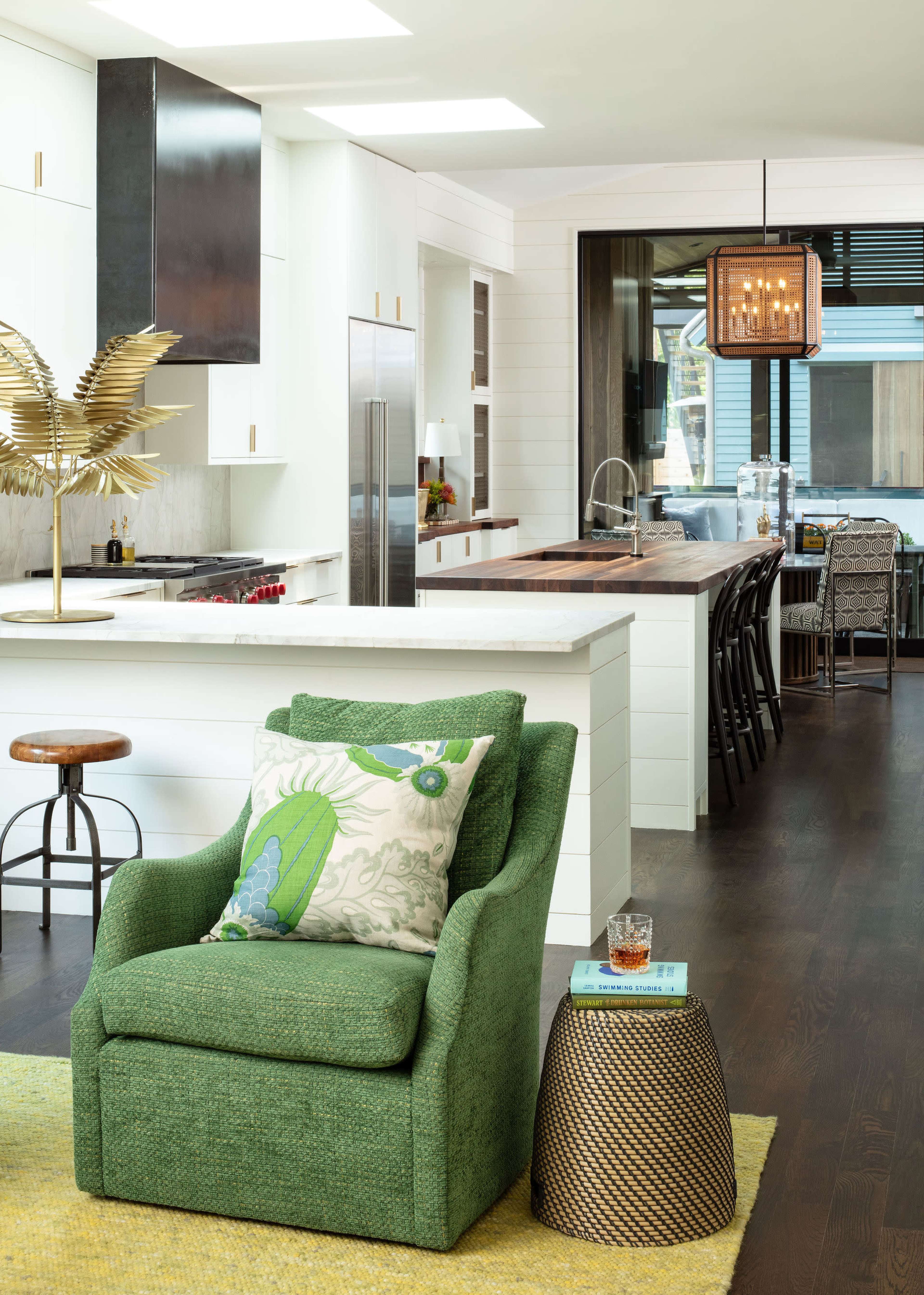 A green upholstered chair with a decorative pillow is positioned next to a small table in a modern kitchen and dining area featuring white cabinets and a large island.