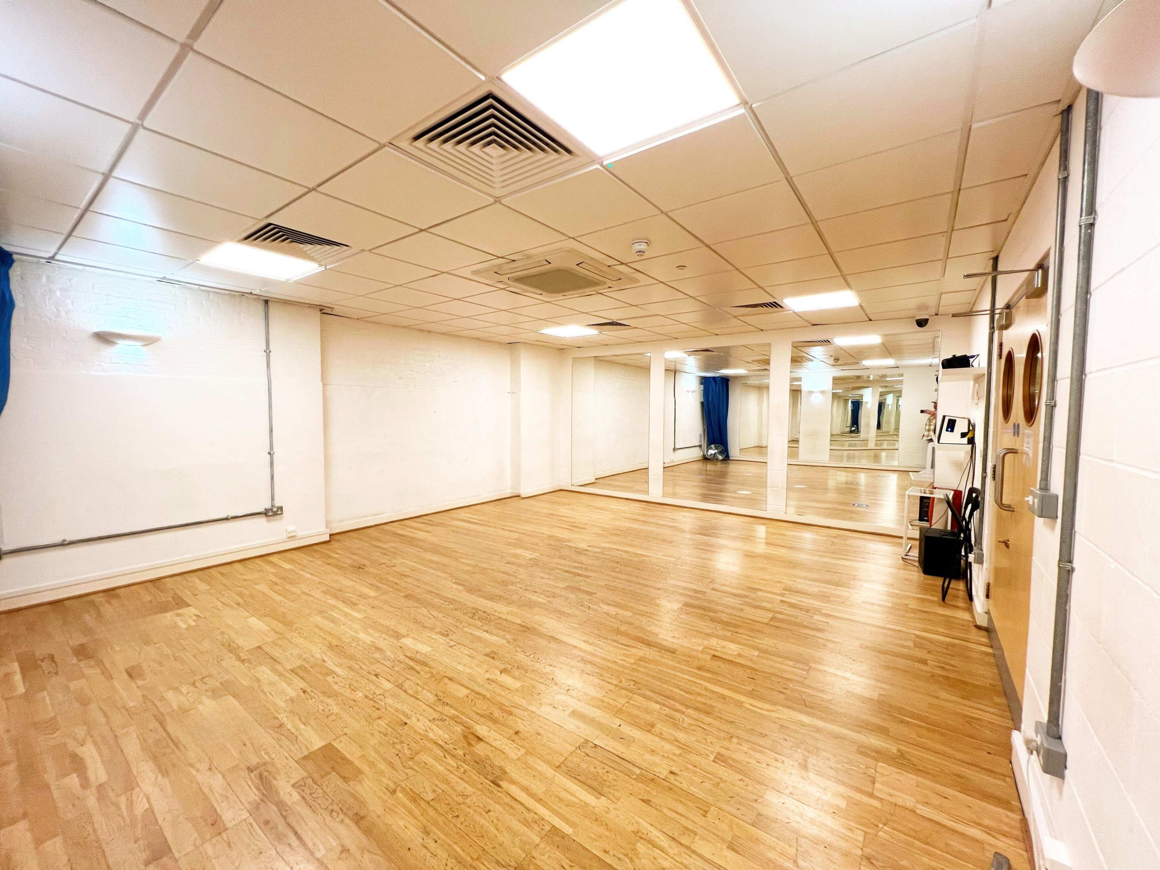 The image shows a spacious, empty studio with wooden flooring, bright overhead lights, and a large mirror along one wall.