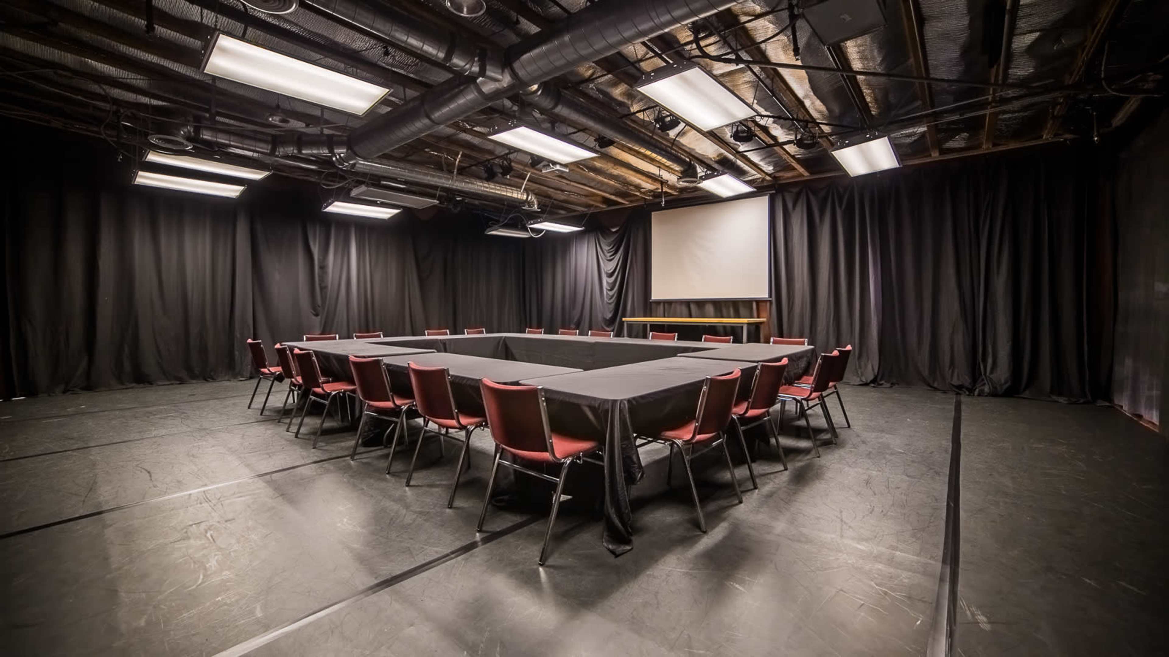 The image shows a large meeting room with a rectangular table surrounded by red chairs, set against black curtains and a projector screen.