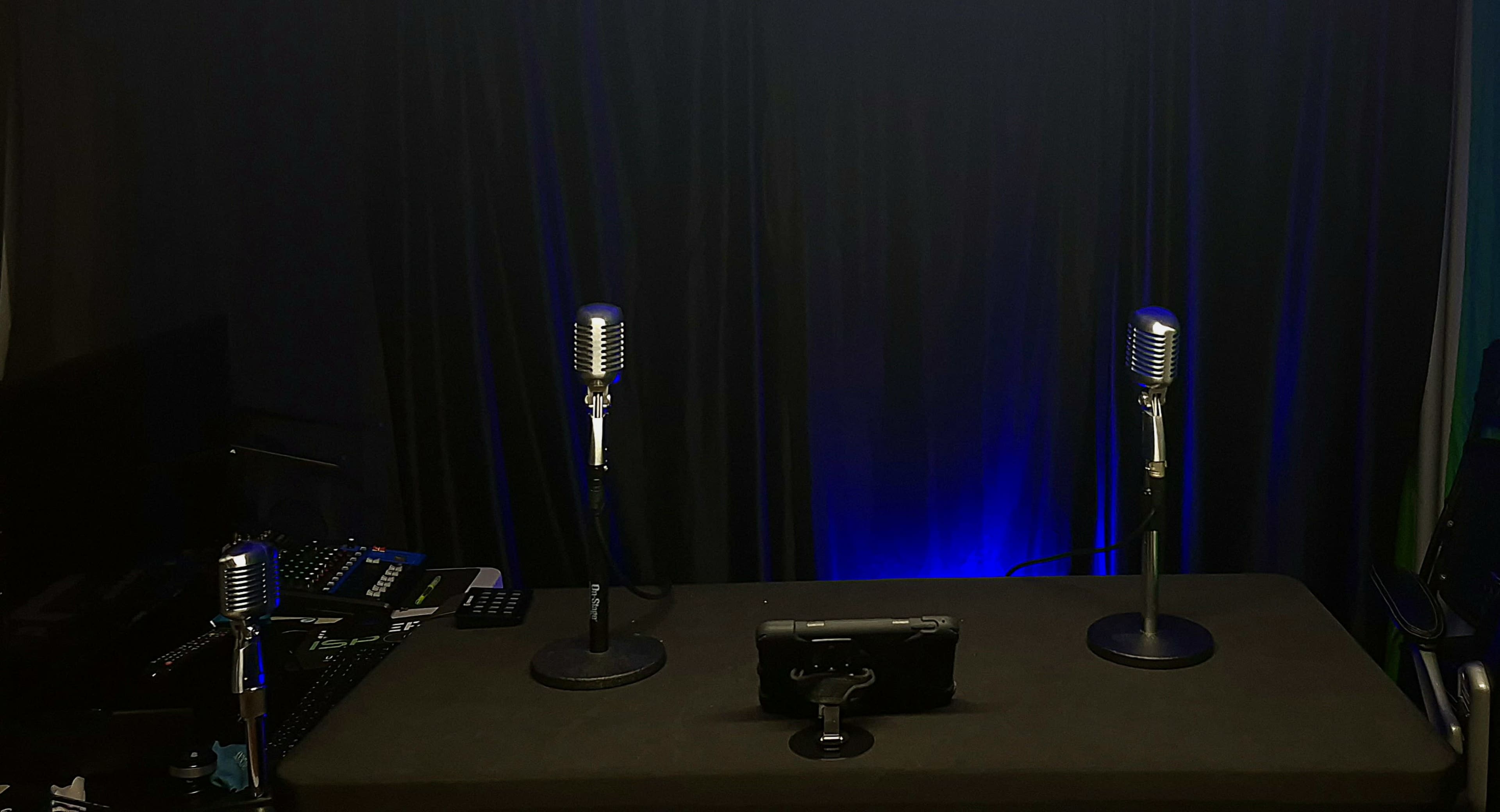 The image shows a dimly lit setup with three microphones on stands positioned at a table with a tablet in the center, against a black curtain backdrop.