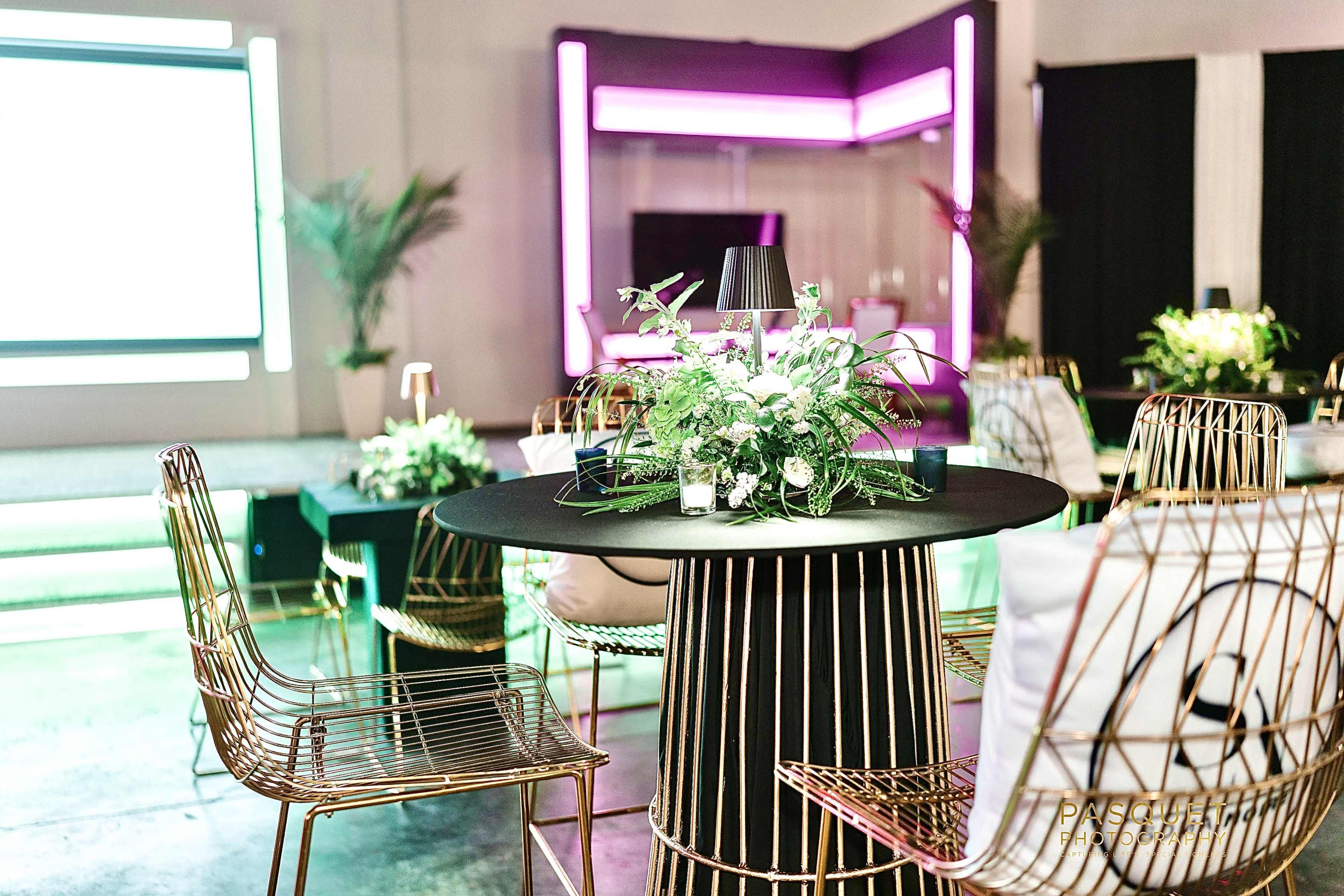 The image shows a modern event space with gold wire chairs arranged around a black table, adorned with greenery and illuminated by colorful lights.