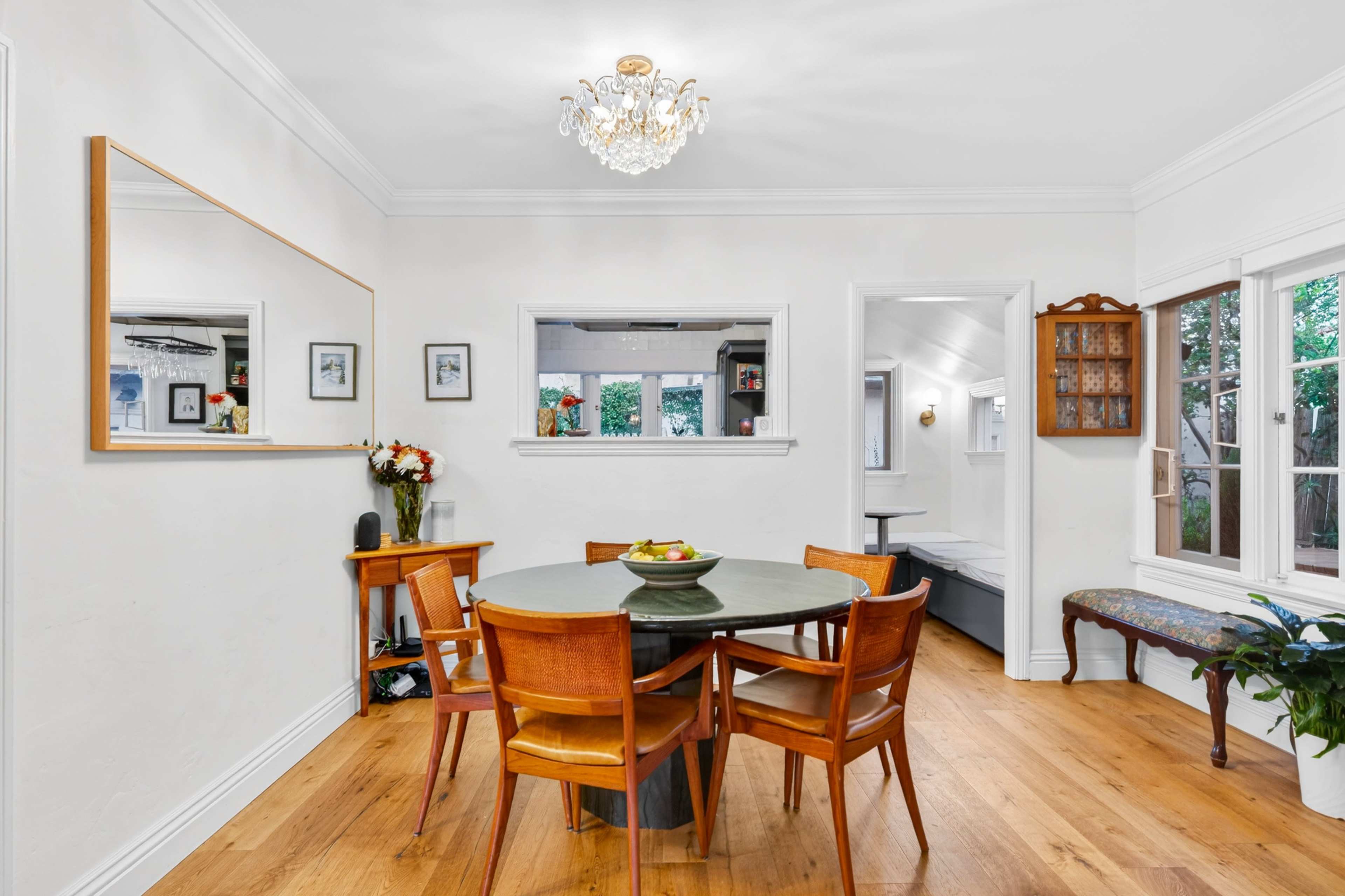 A dining area features a round table surrounded by wooden chairs, a chandelier above, and a large mirror reflecting the bright, open space.