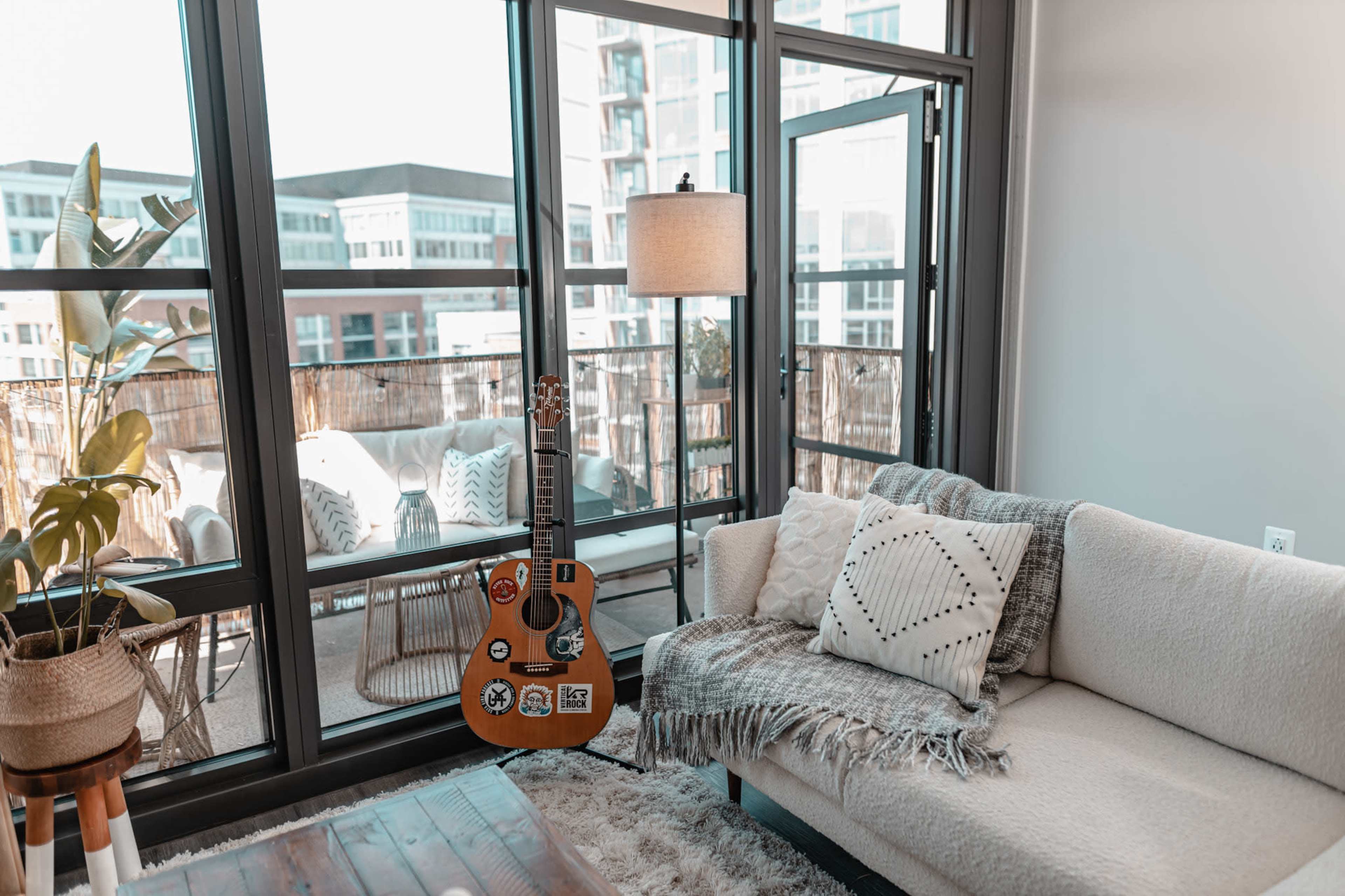 A guitar with stickers leans against a light-colored couch near large windows overlooking a balcony with outdoor seating.