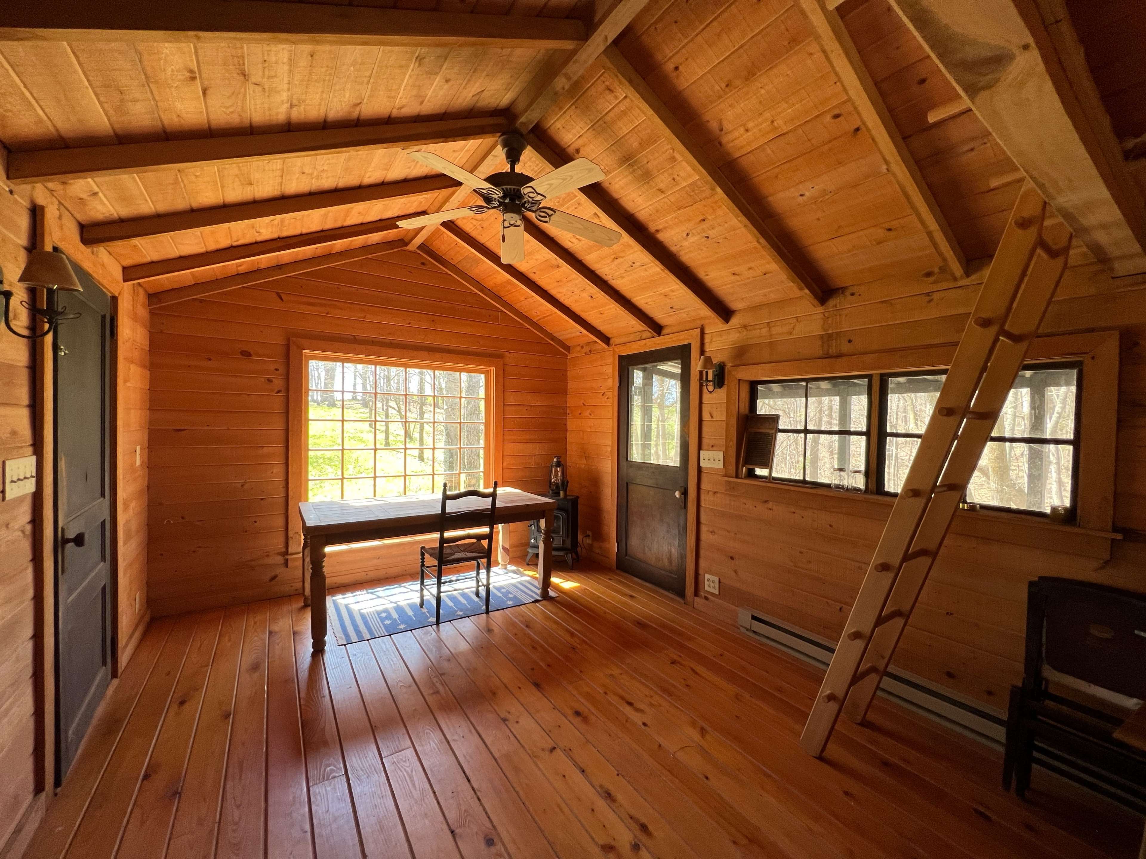 The interior of a wooden cabin features a table and chairs near a large window, with a ladder leading to a loft area.