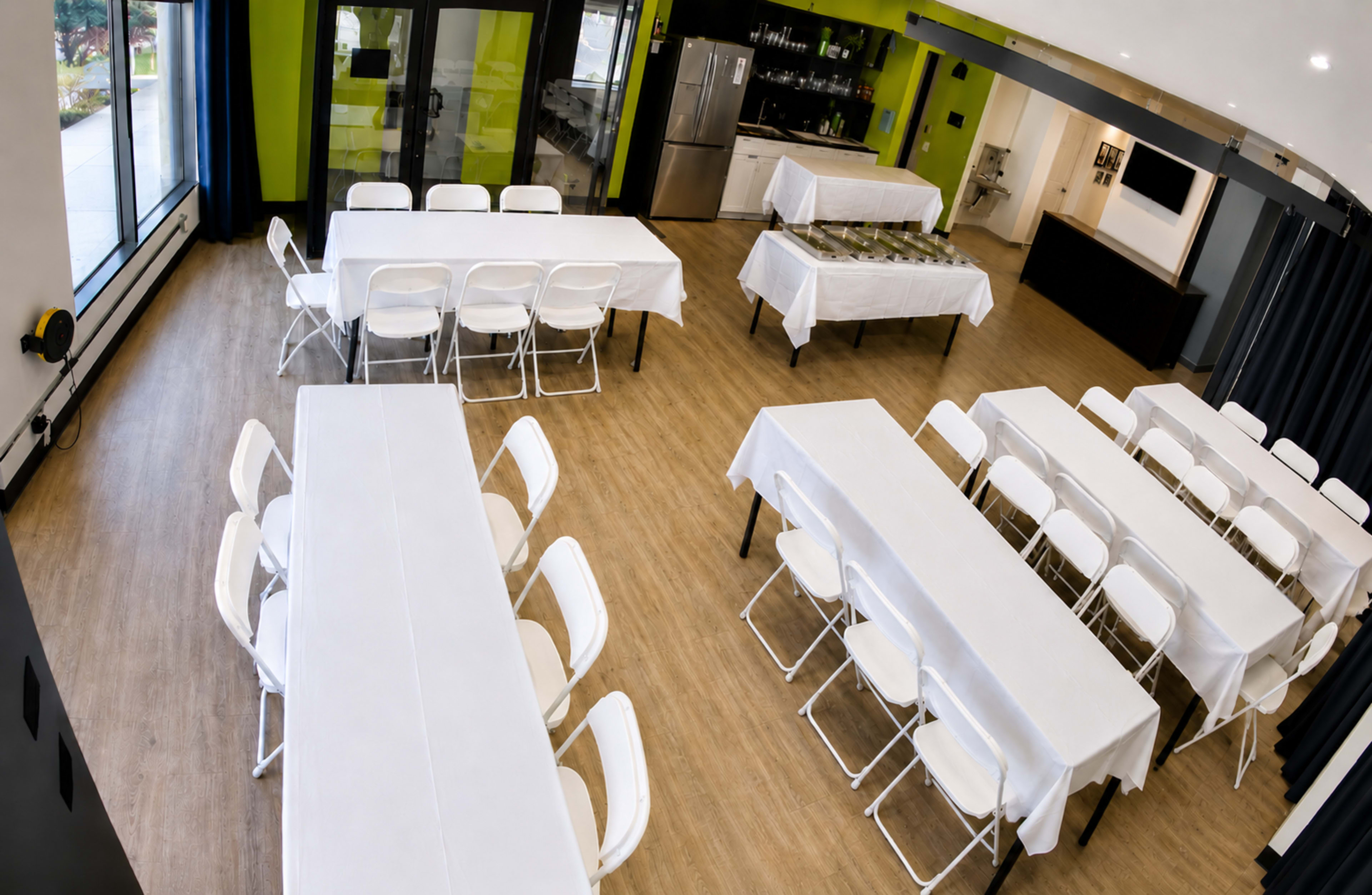 The image shows a spacious indoor setting with multiple white tables and chairs arranged neatly on a wooden floor, featuring a serving table with food at the back.