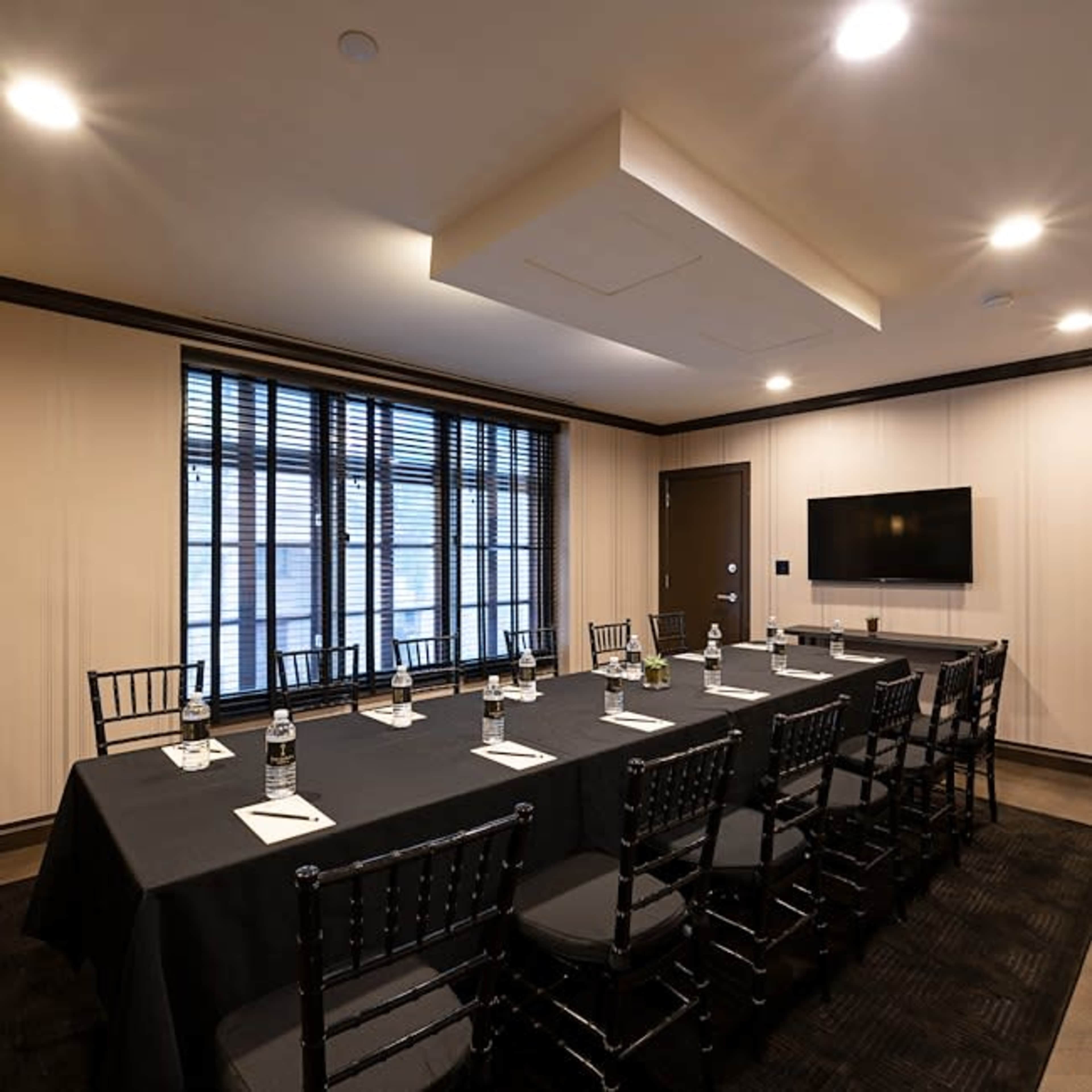 A conference room features a long black table surrounded by black chairs, with a TV mounted on the wall and large windows covered by blinds.
