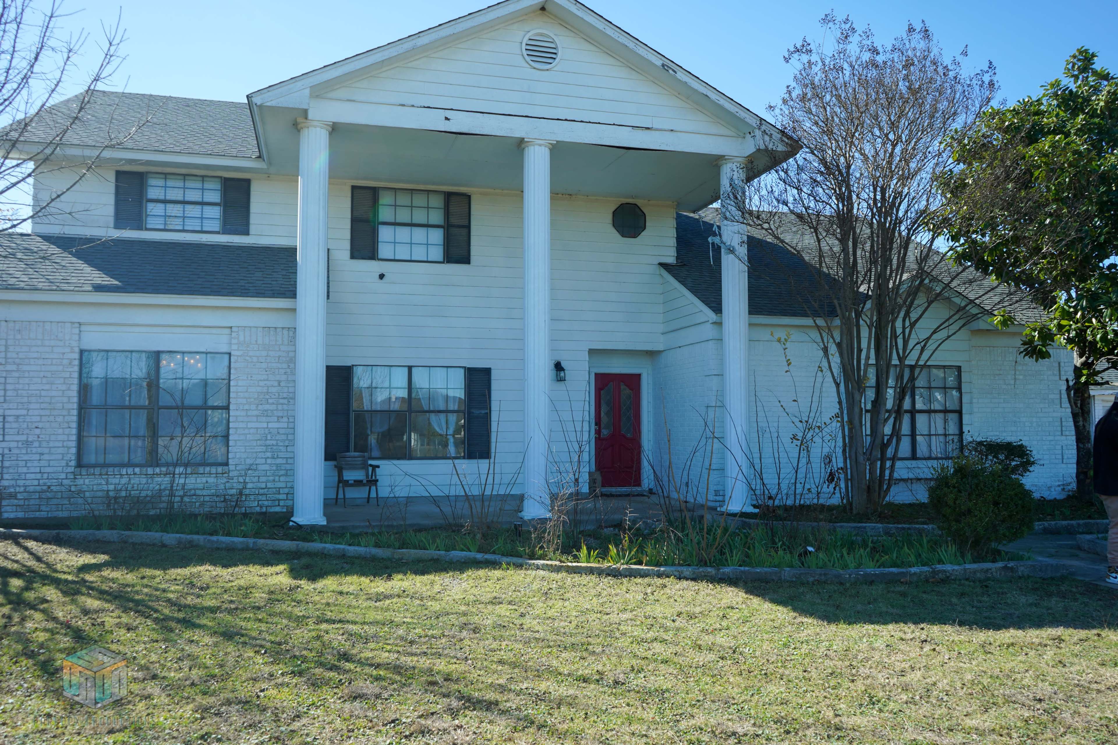 A two-story house features four columns supporting the front porch and a red door, with a yard of sparse vegetation.