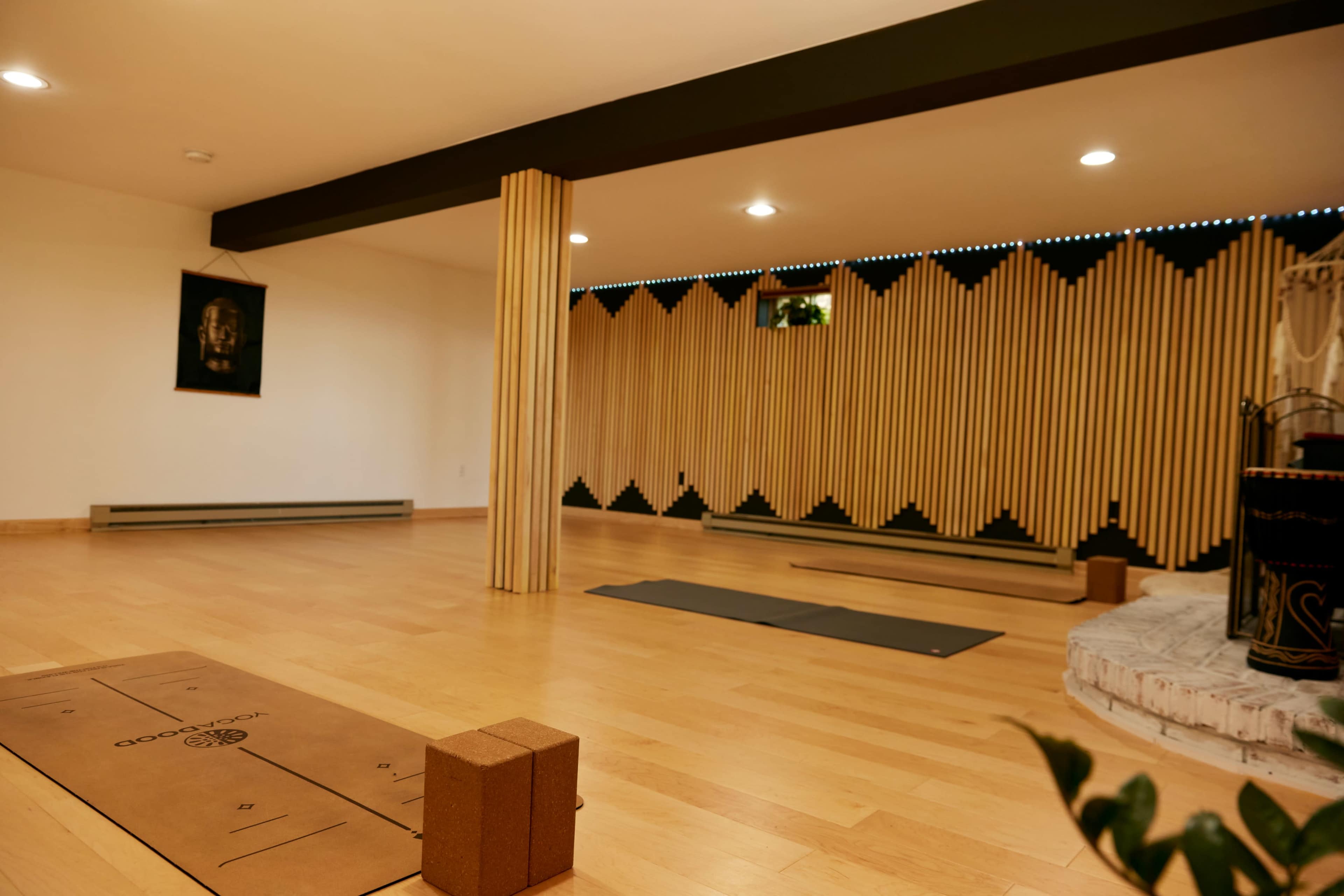 A spacious yoga studio featuring wooden floors, a decorative wall with vertical wooden elements, and yoga mats arranged on the floor alongside props.