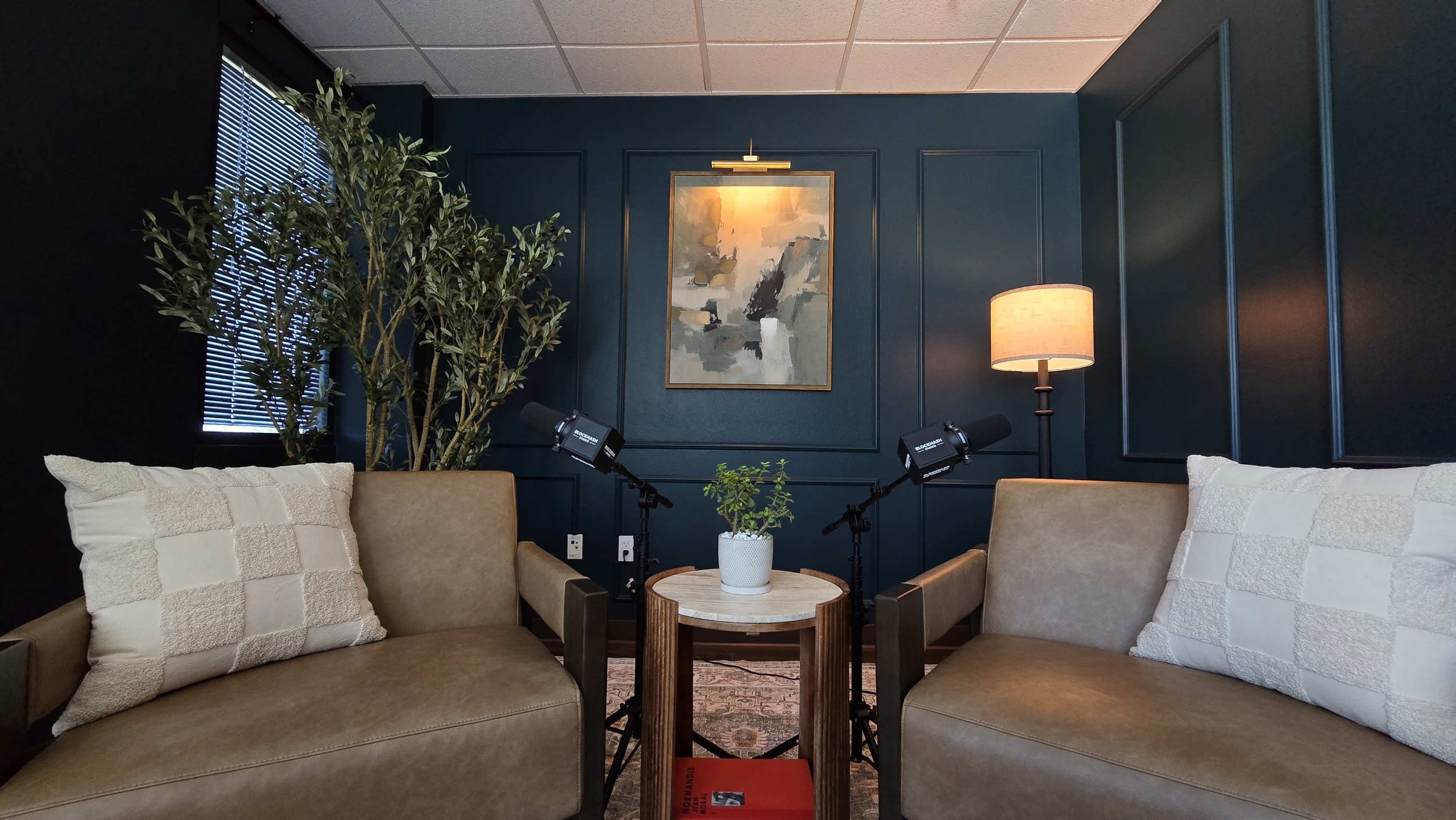 The image shows a small room with two light brown armchairs facing a round table, a potted plant on it, artwork on the wall, and a lamp beside one chair.