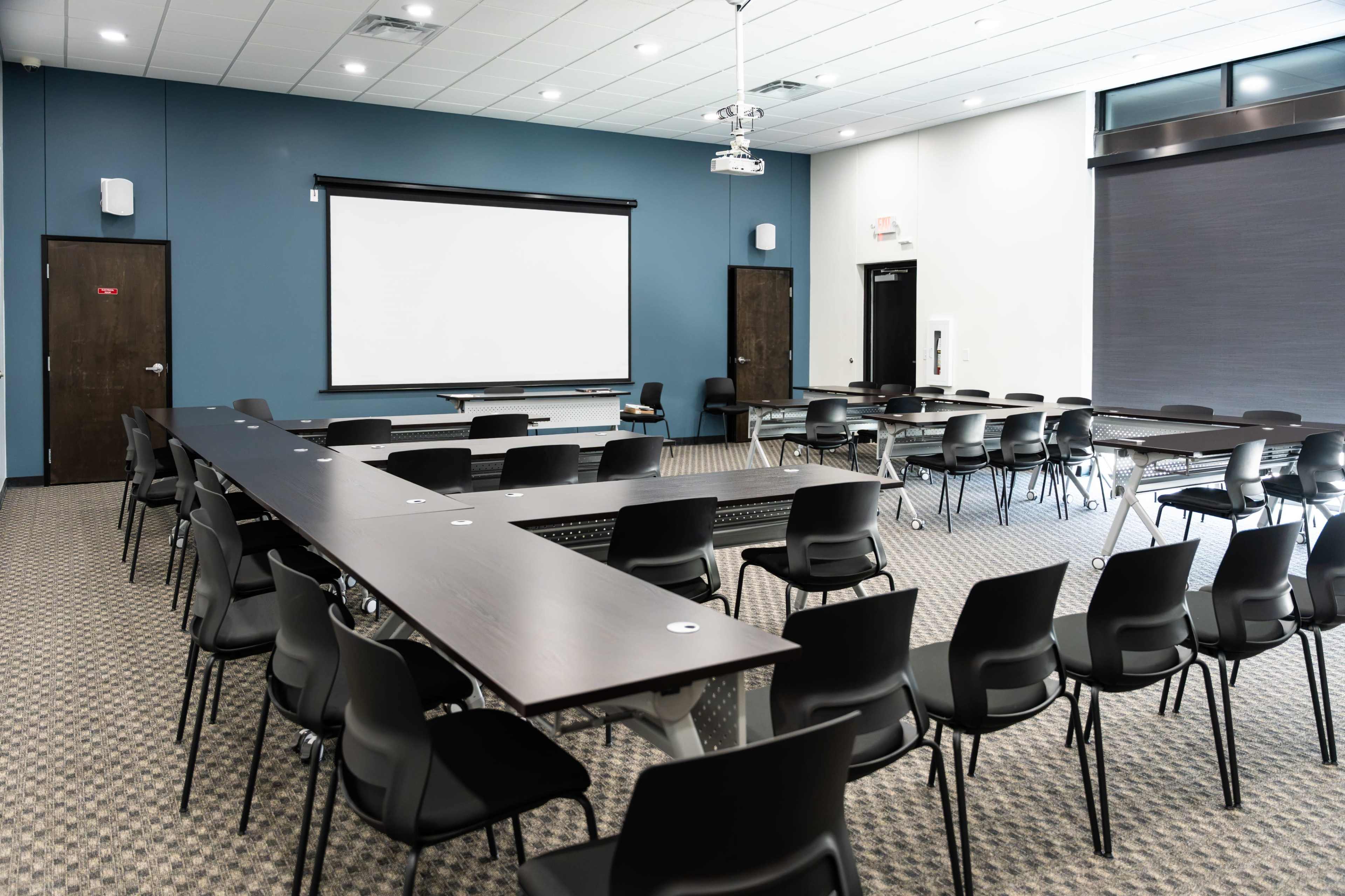 The image shows a modern conference room arranged with several black chairs and long tables facing a large screen at the front.