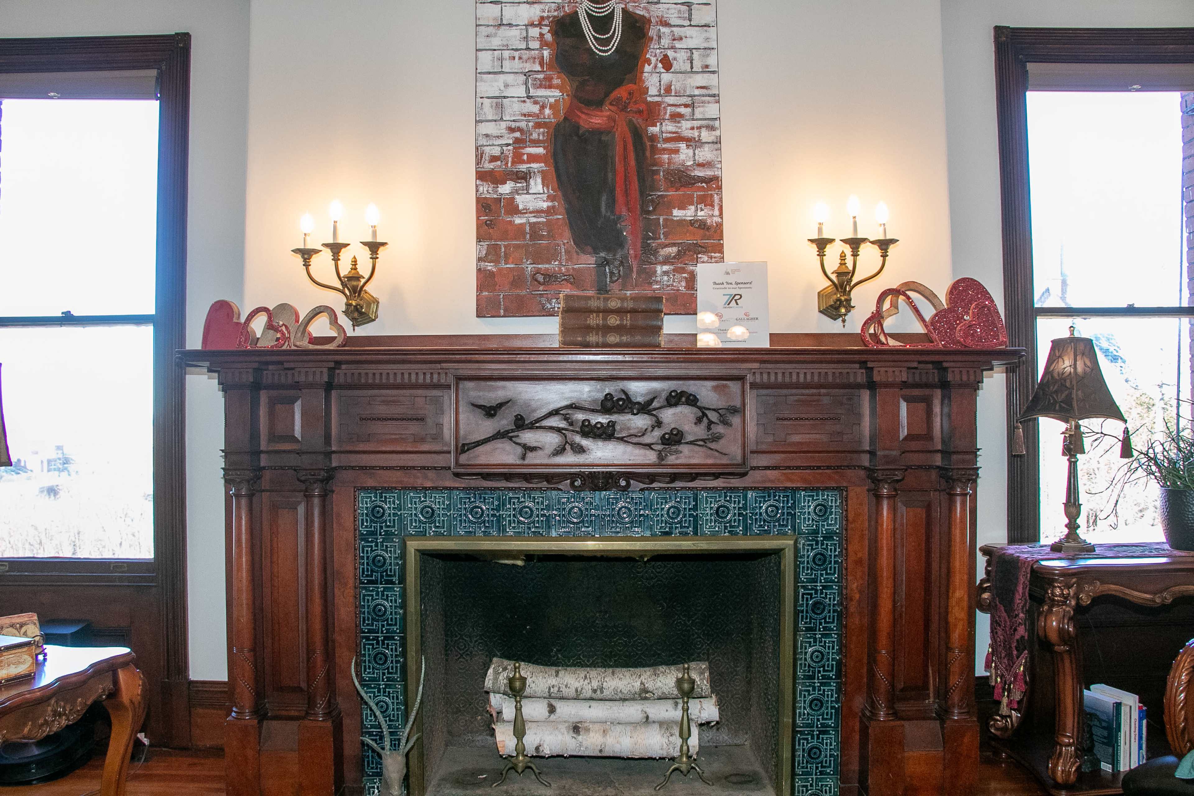 A decorated fireplace mantel with a painting of a figure dressed in black, flanked by candelabra and heart-shaped decorations.