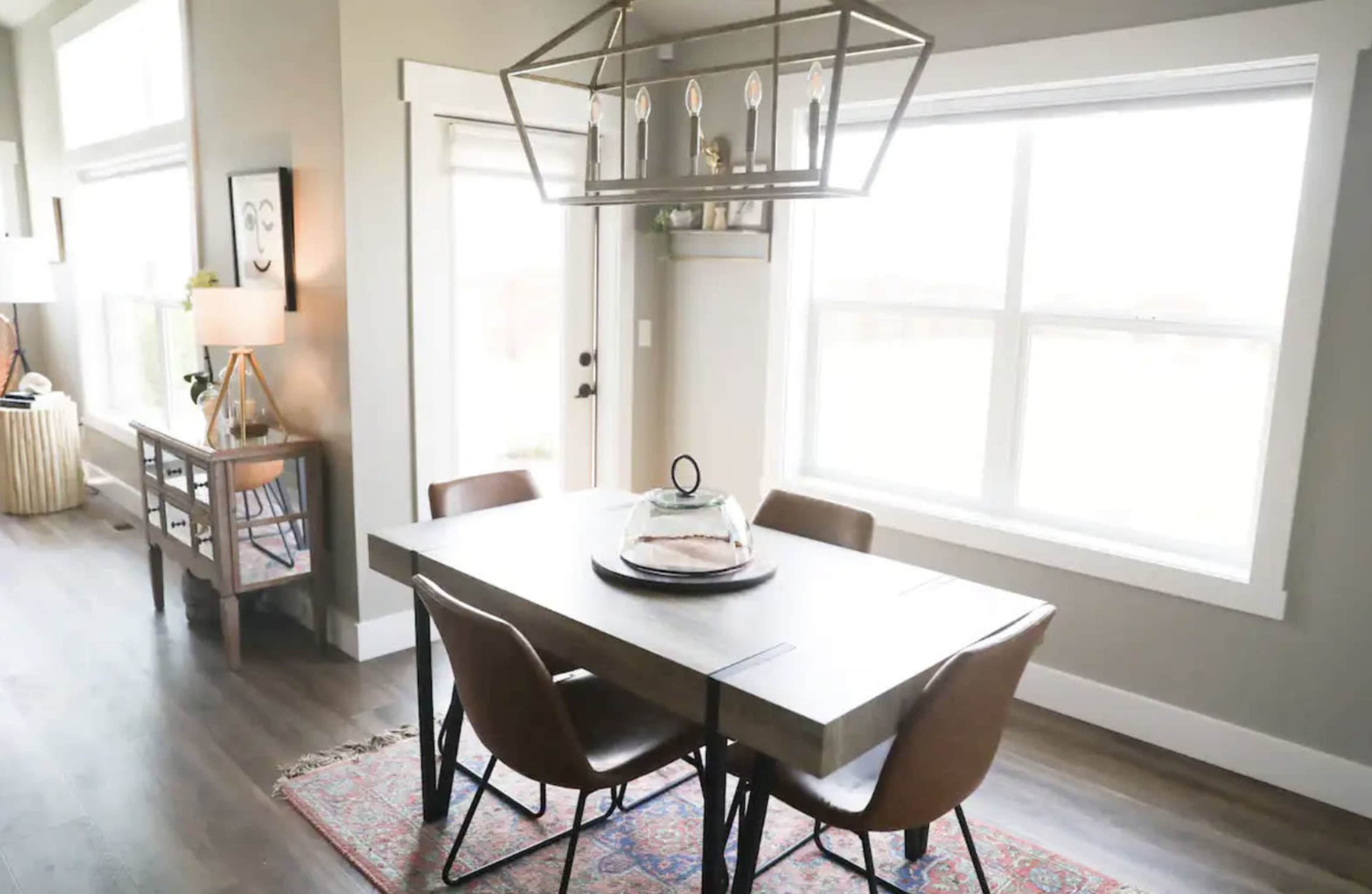 A dining area features a rectangular table with four chairs, a decorative chandelier above, and large windows allowing natural light.