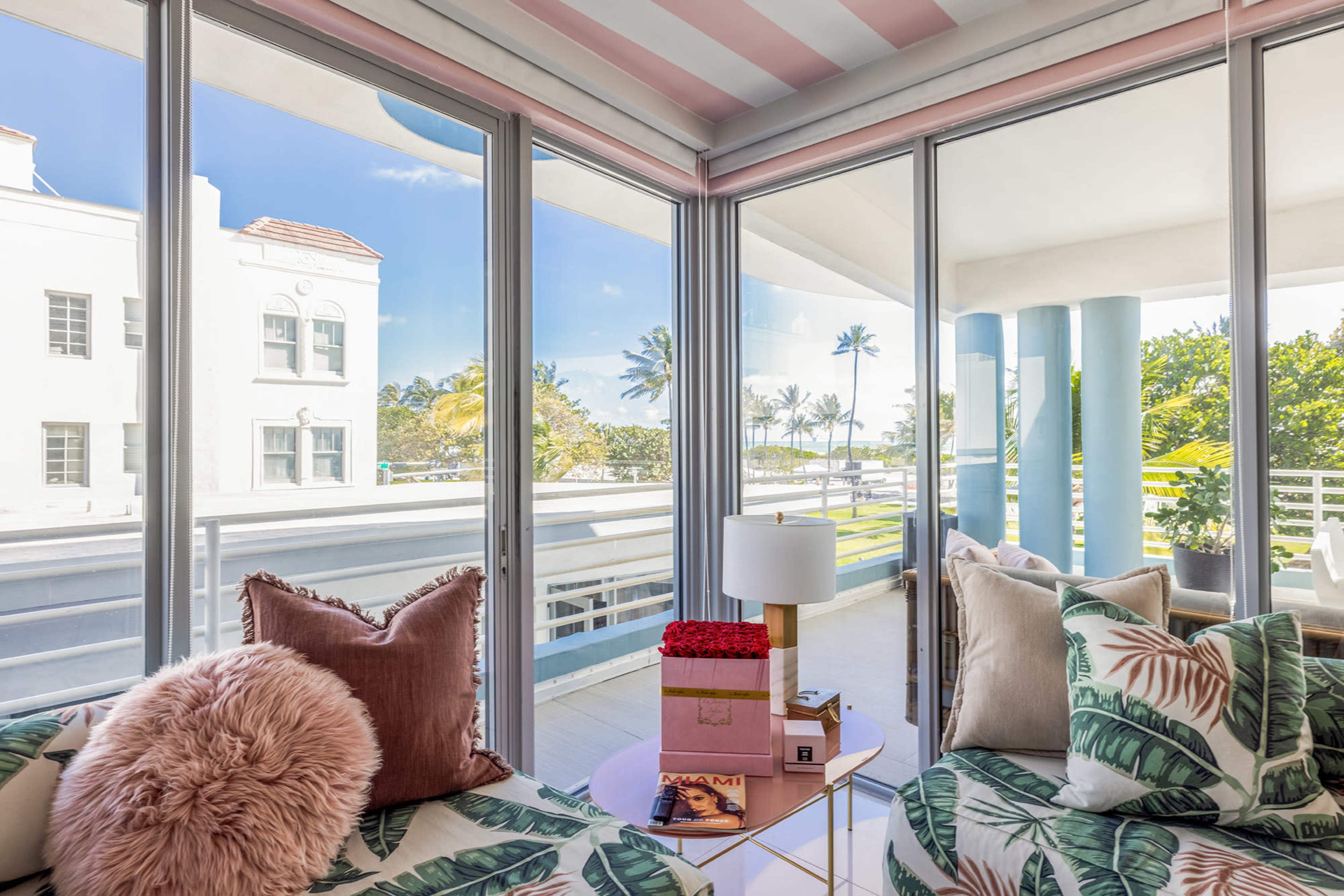 A bright living space features large glass windows overlooking a balcony with palm trees in the background.