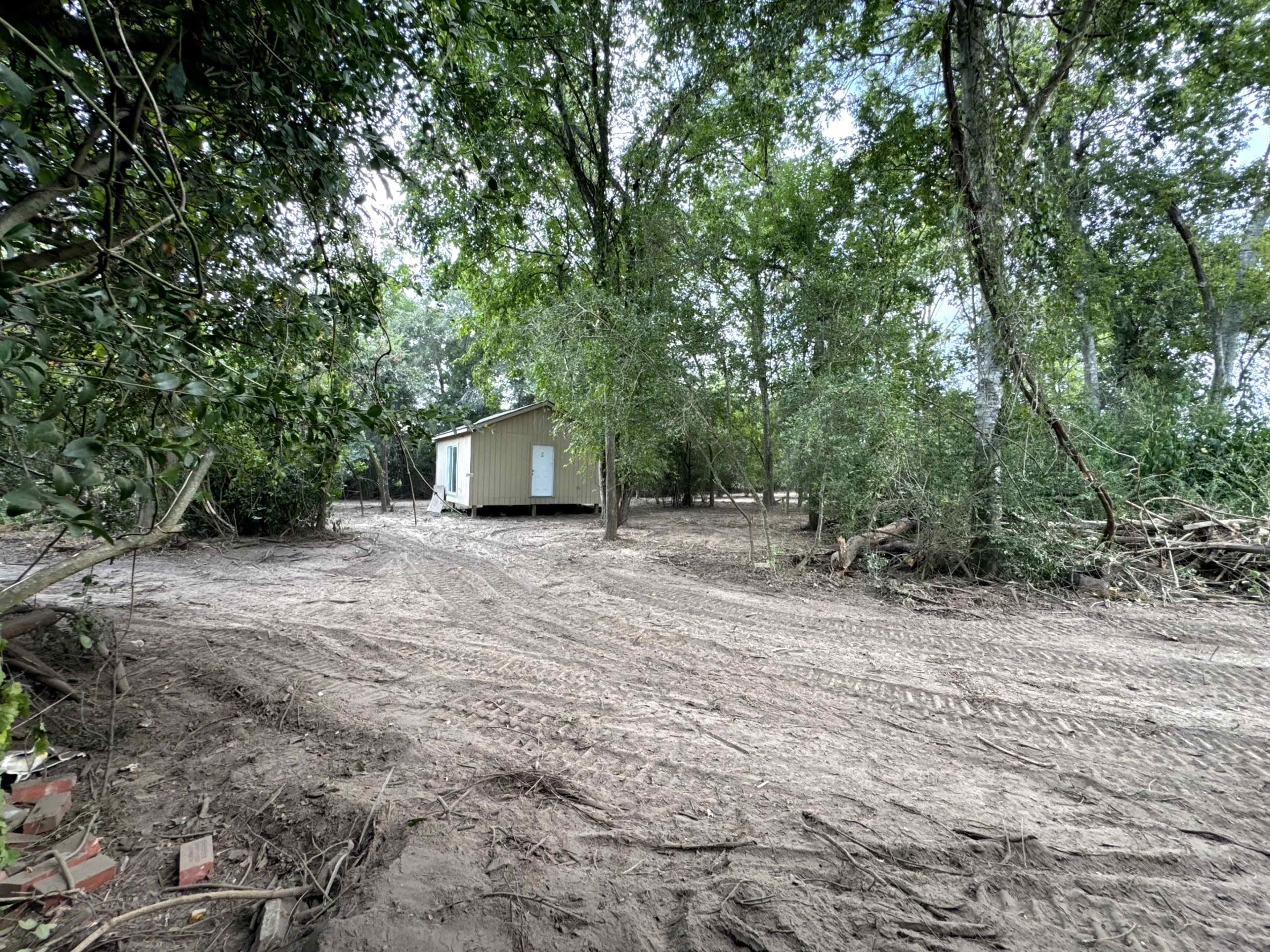 A small, unfinished building sits amidst a clearing in a wooded area, surrounded by trees and exposed earth.