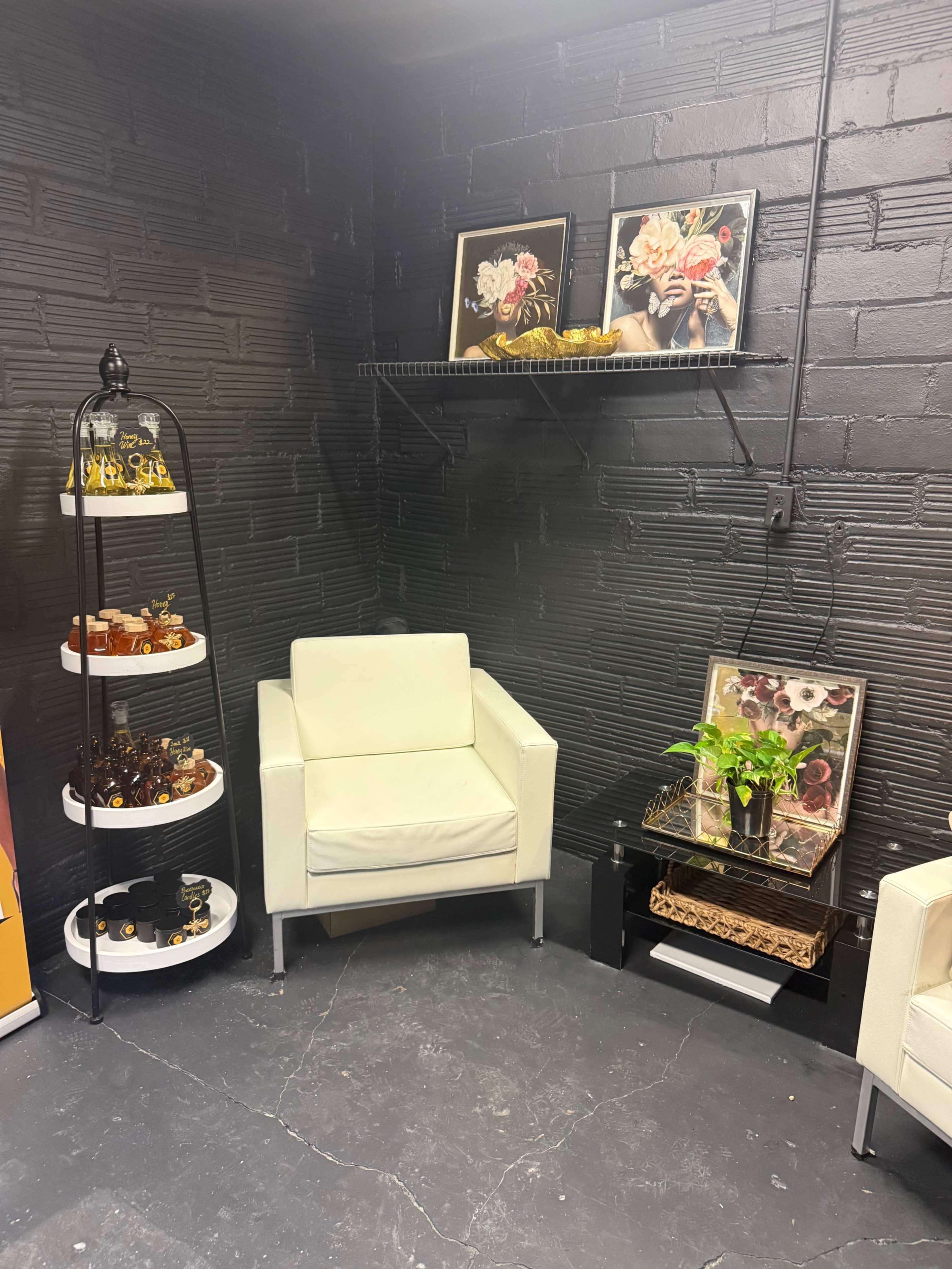 The image shows a corner of a room featuring a white armchair, a small table with a decorative plant, and shelves displaying various items, all set against a black textured wall.