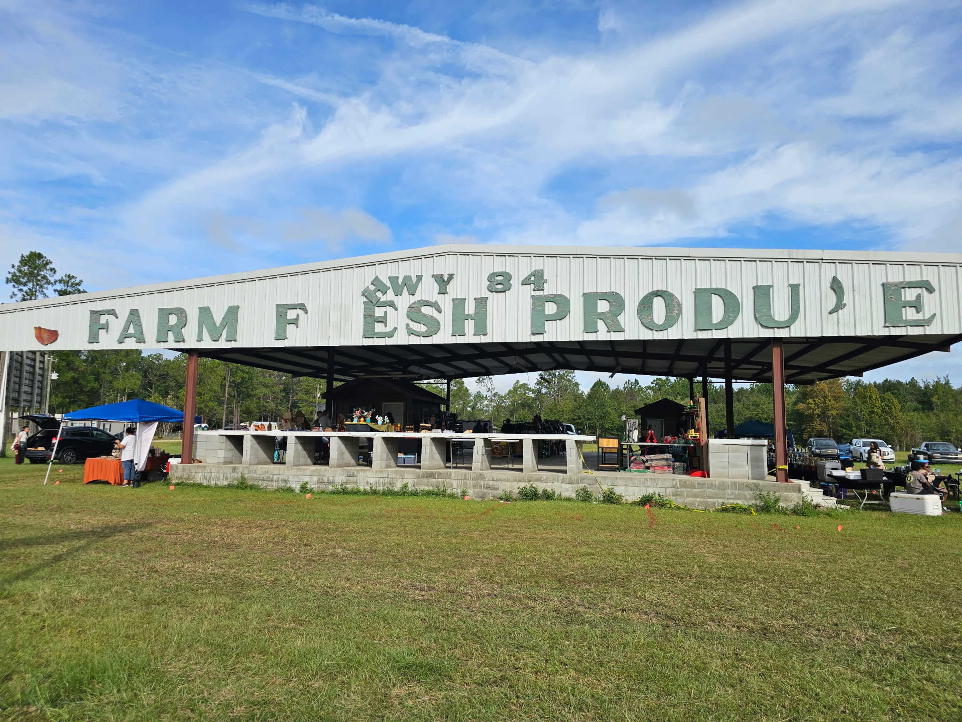 The image shows a large metal barn with a sign reading "FARM FRESH PRODUCE" under a clear blue sky, surrounded by green grass and various tents.