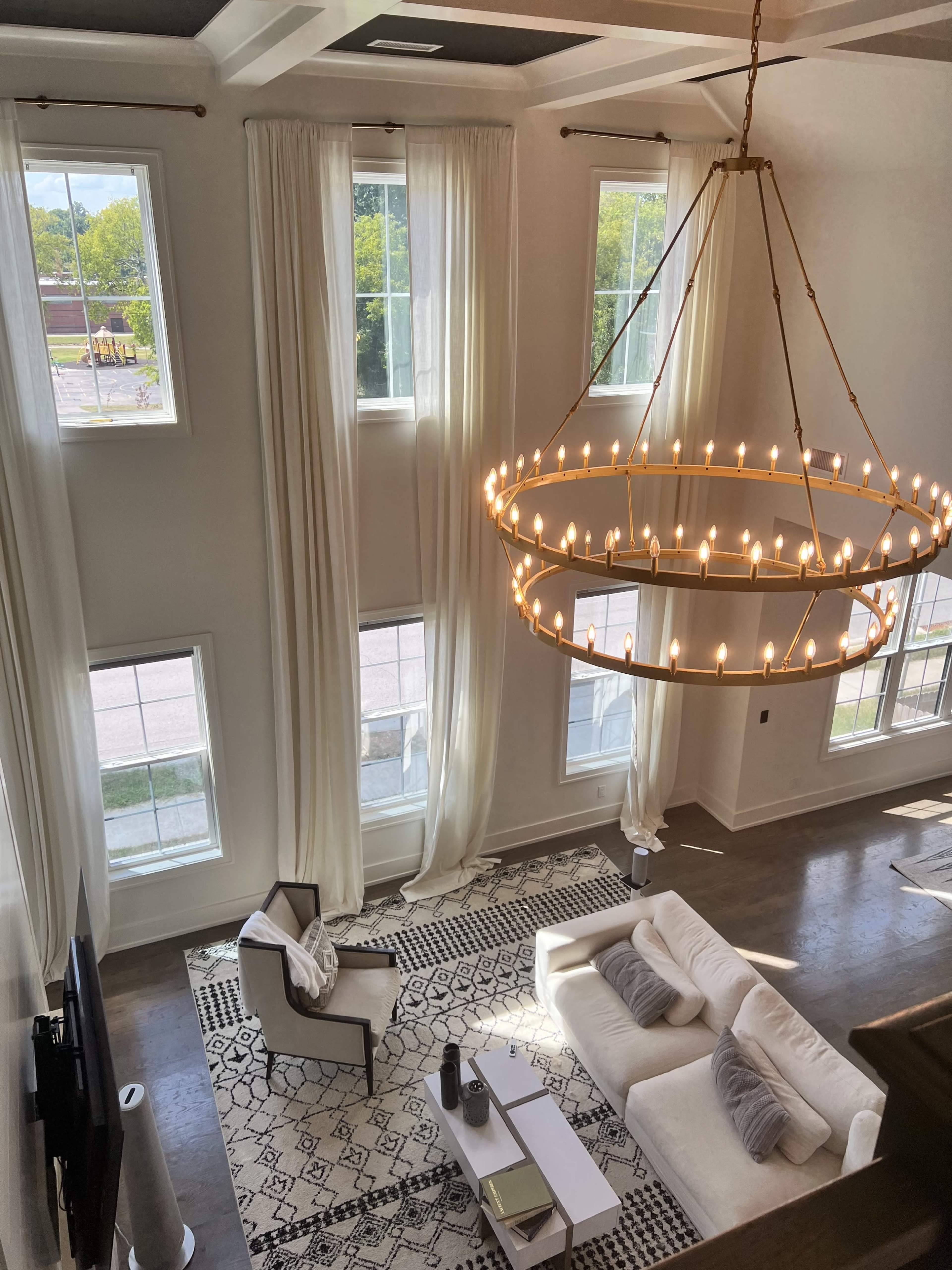 A spacious living room with large windows, a modern chandelier, and a white seating arrangement on a patterned rug.