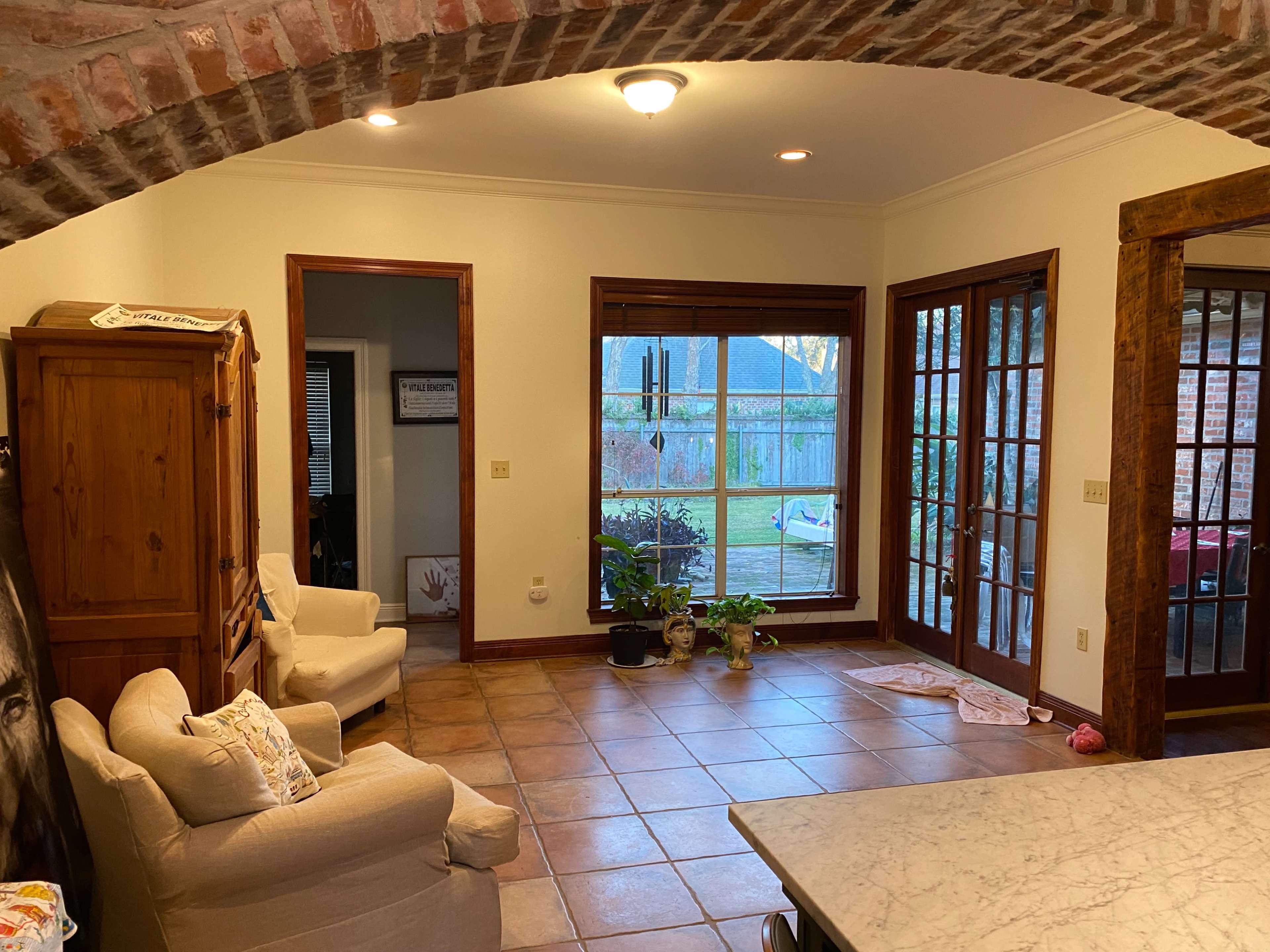 A kitchen with a tiled floor, a brick archway, an armchair, and a large window providing a view of a backyard.