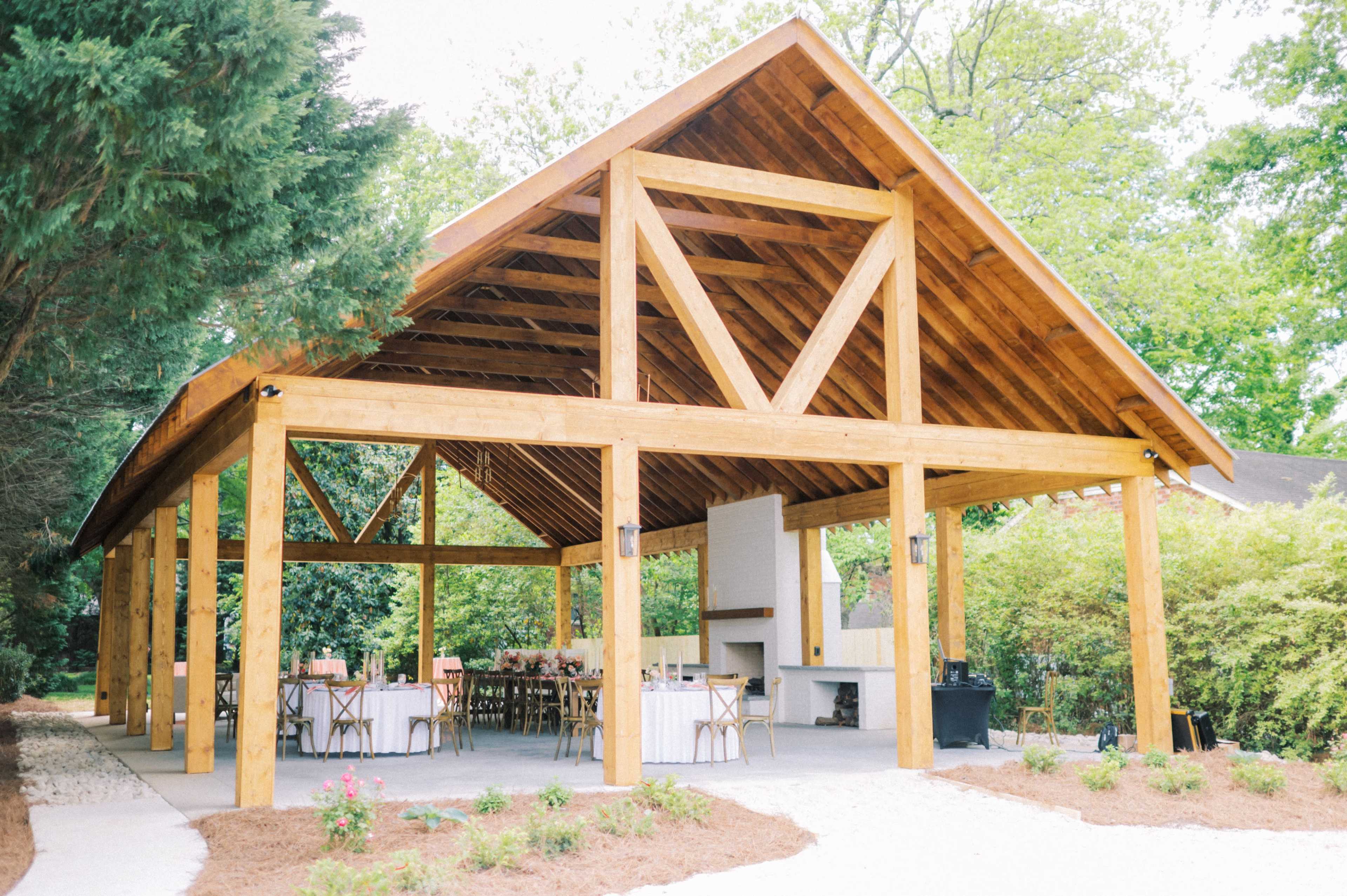 The image shows a wooden pavilion with a sloped roof and open sides, featuring tables arranged for dining under the structure.