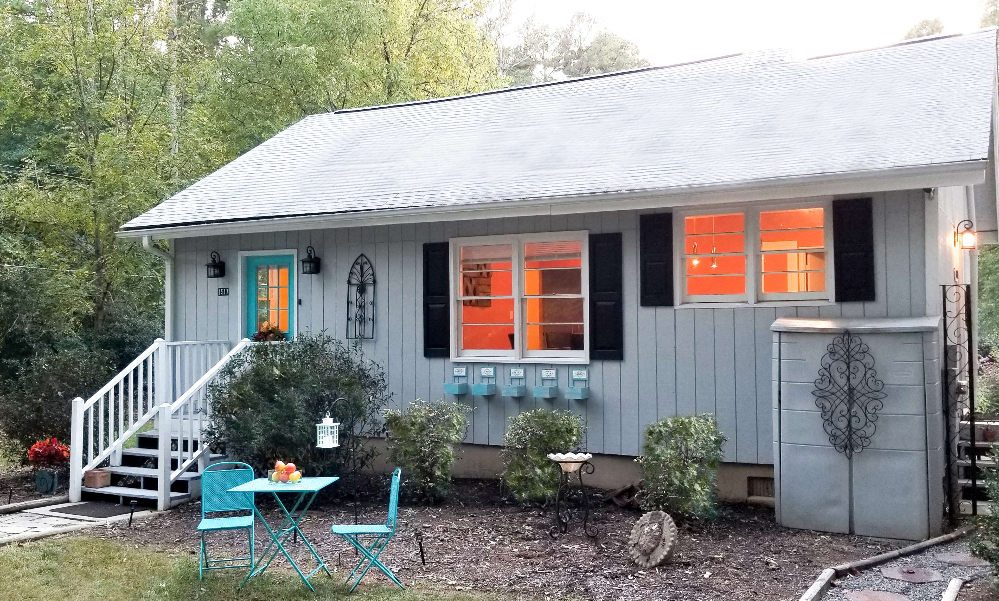 A small, gray house features a turquoise door and shutters, with a set of mailboxes and a blue table and chairs in the front yard.