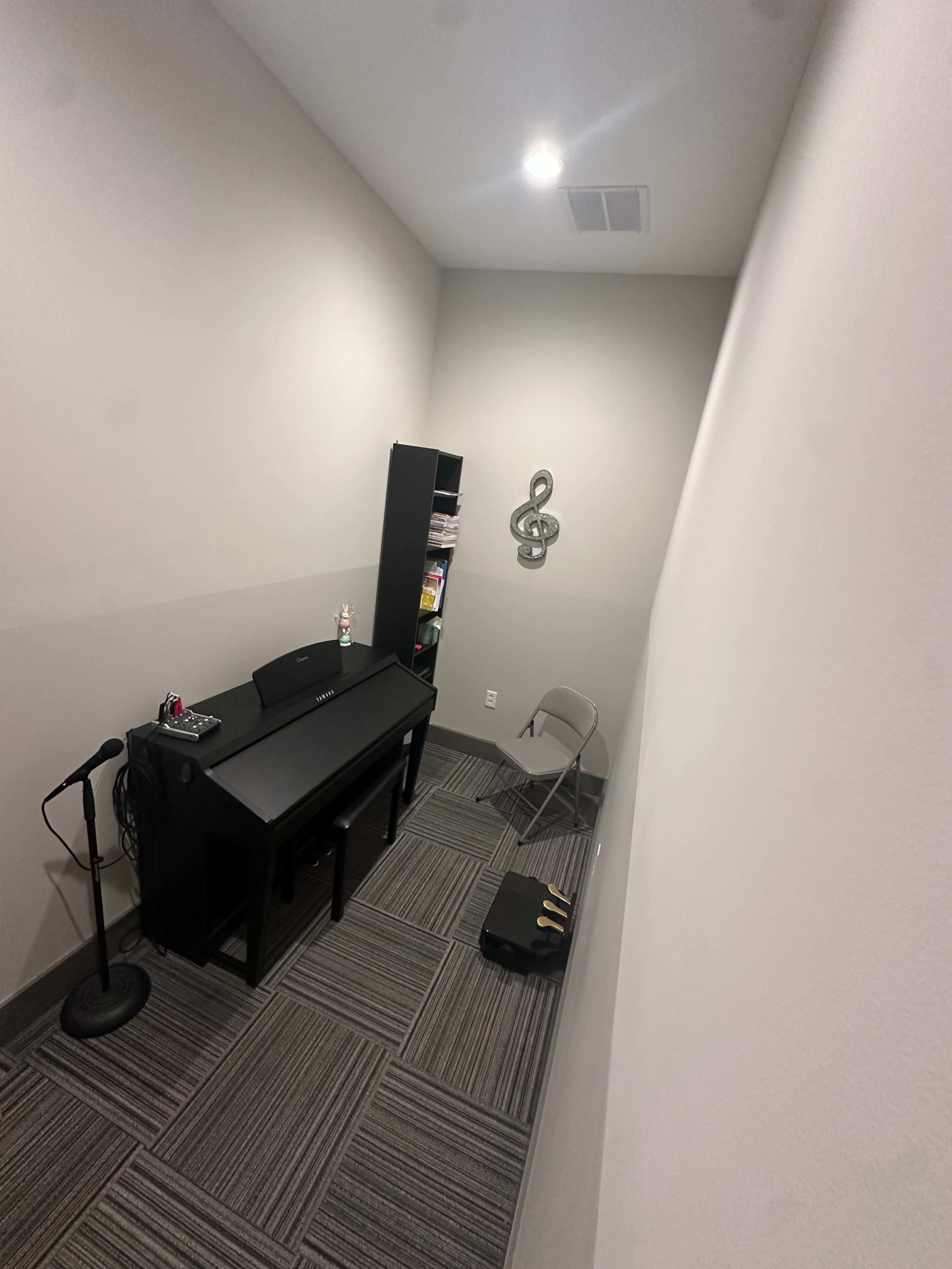 The image shows a small music practice room containing a keyboard, a microphone stand, a chair, and a shelf filled with music books.