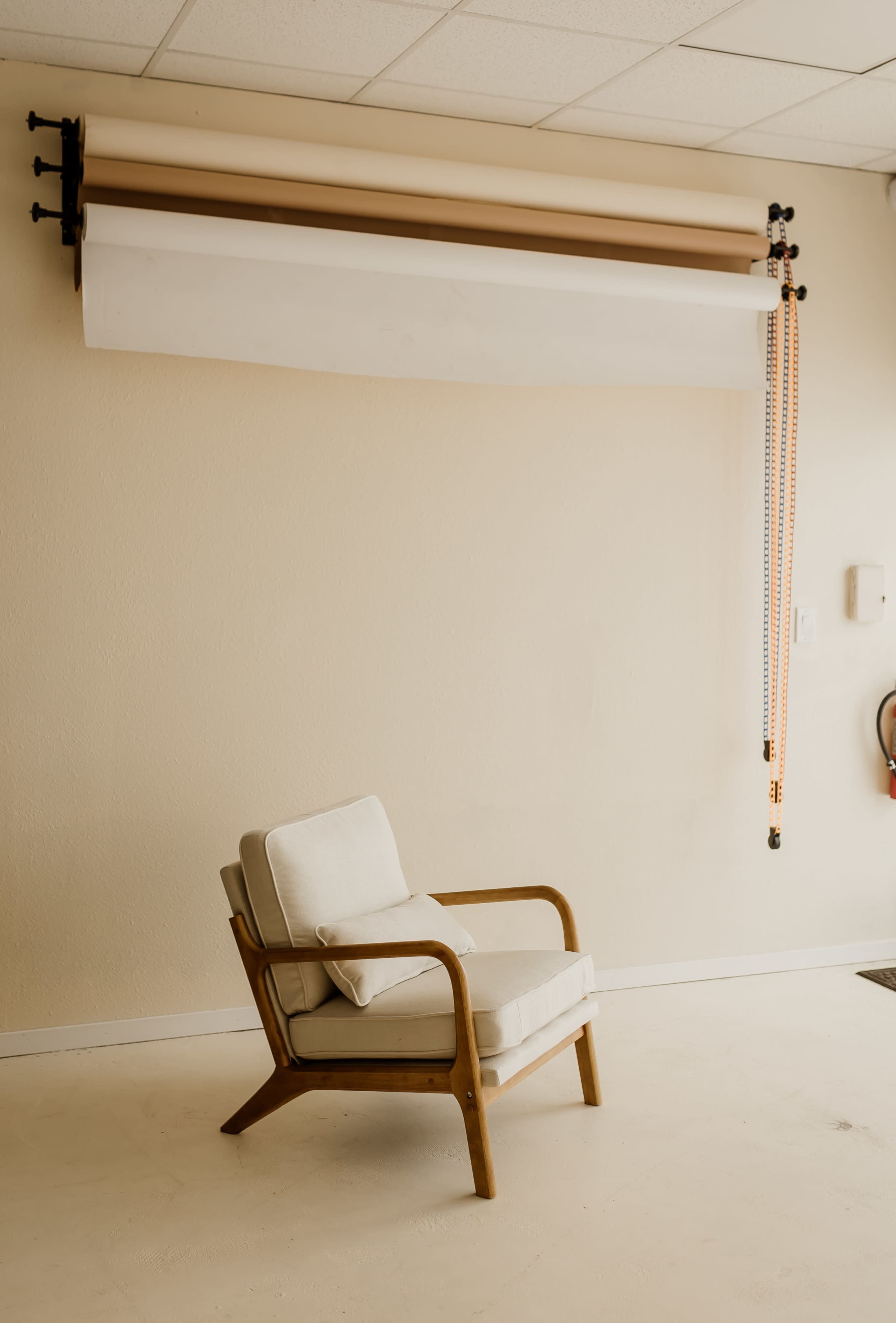A light-colored chair sits against a plain wall with a paper backdrop and hanging cords above it.