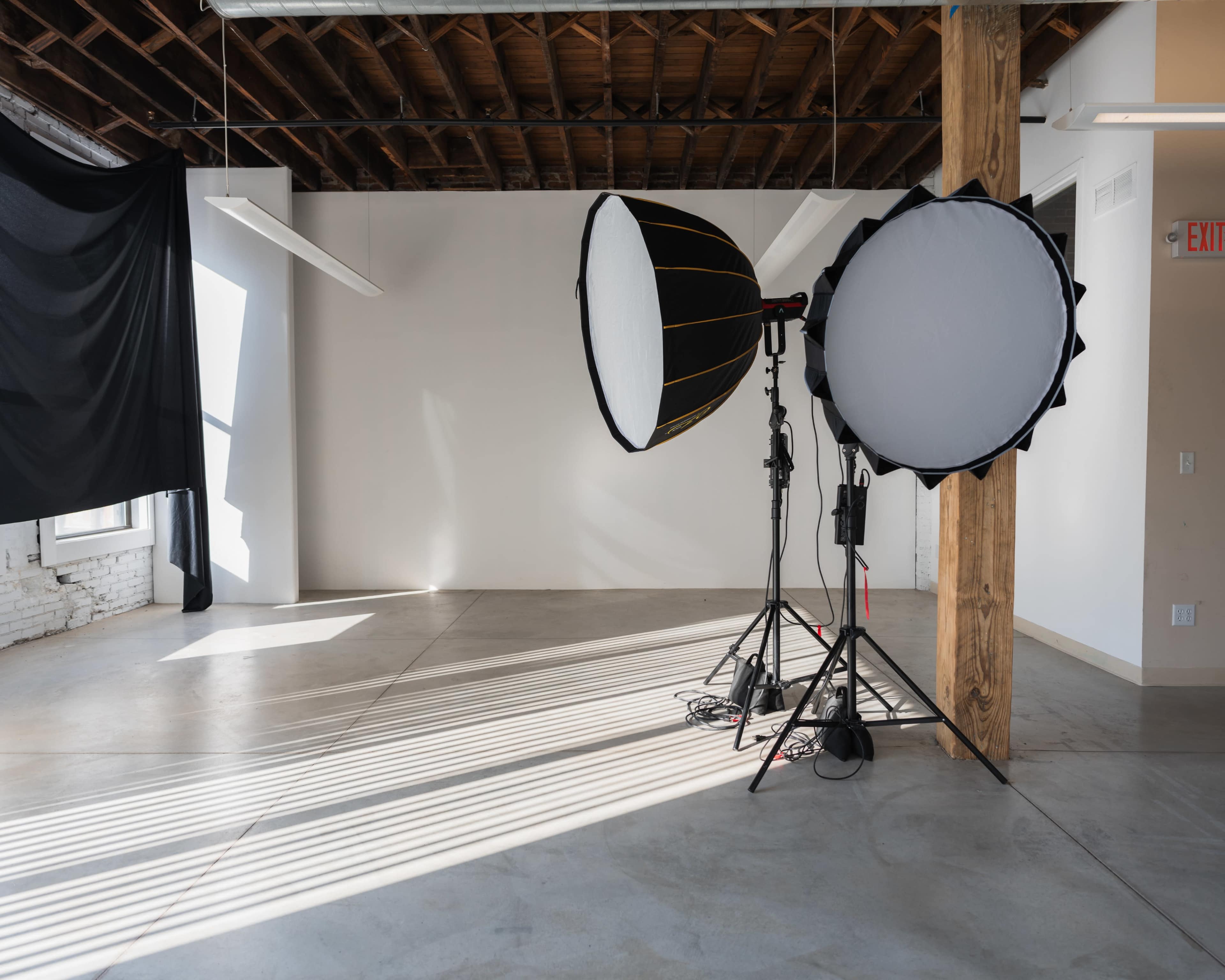 The image shows a photography studio with large softbox lights, a black fabric backdrop, and concrete flooring under natural light.