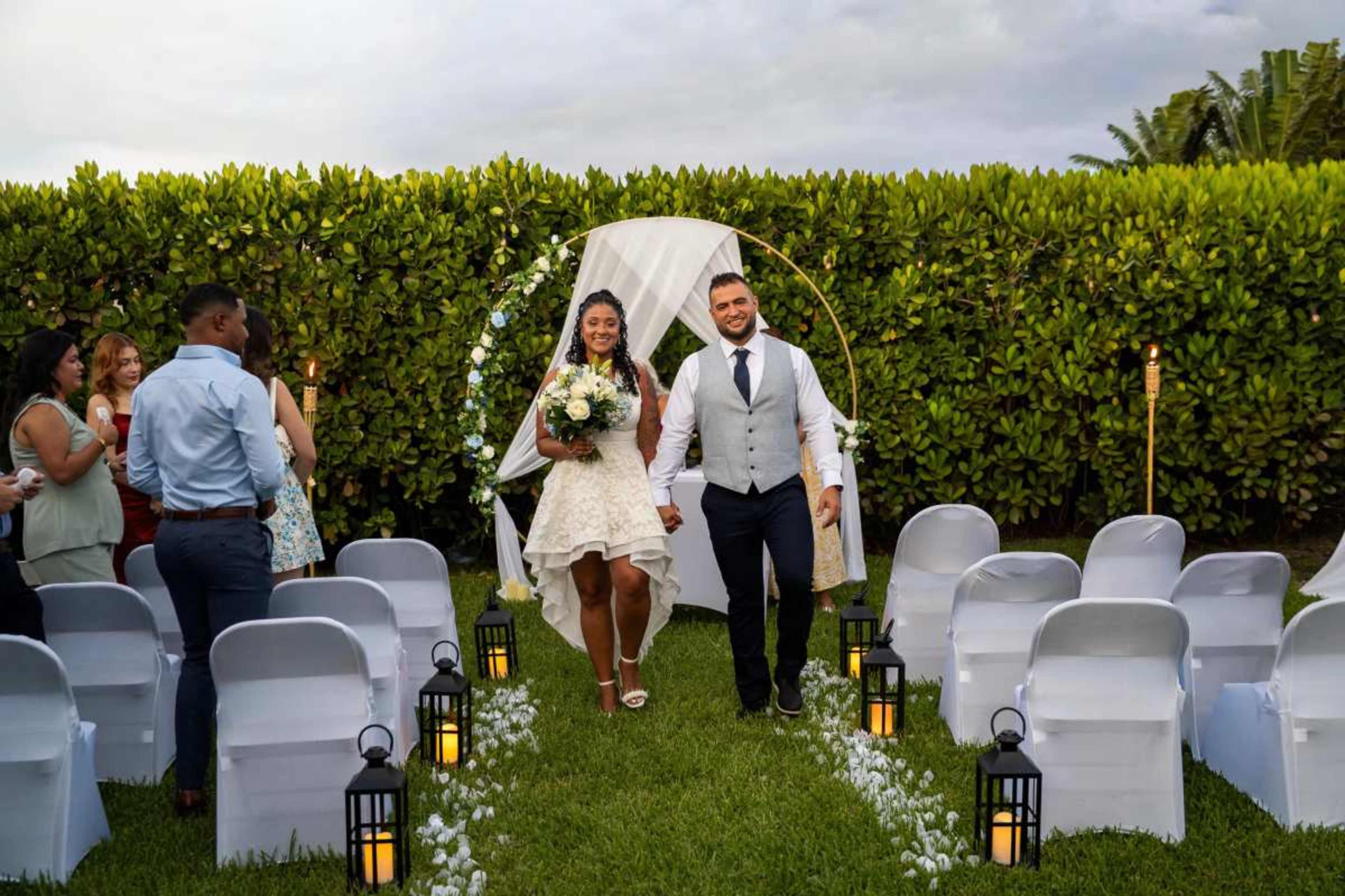 A bride and groom walk down an aisle of white chairs, surrounded by guests and greenery, during an outdoor wedding ceremony.
